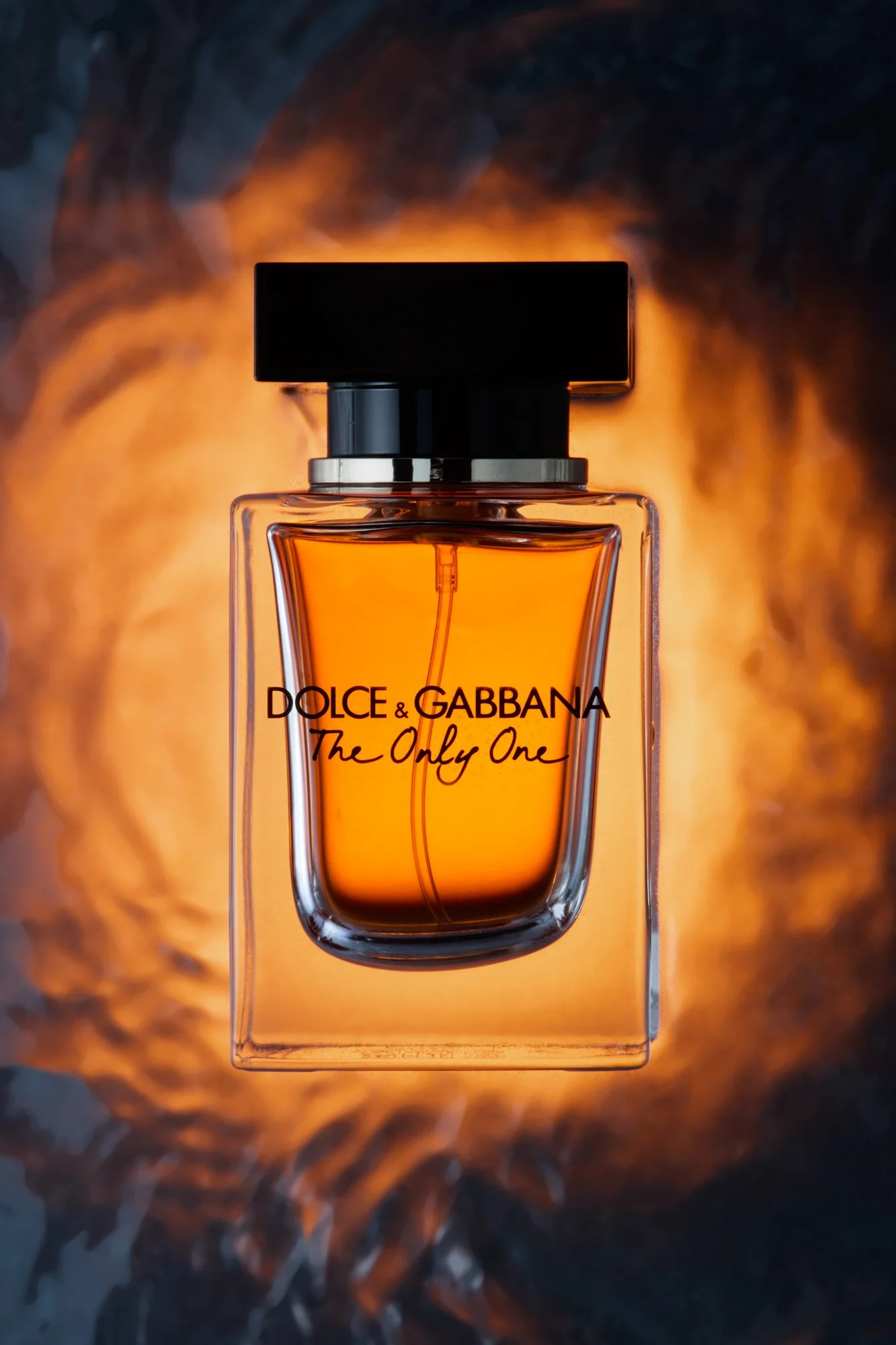 A Dolce & Gabbana perfume bottle labeled 'The Only One' with an orange liquid inside, set against a blurred, dark background with warm orange light.