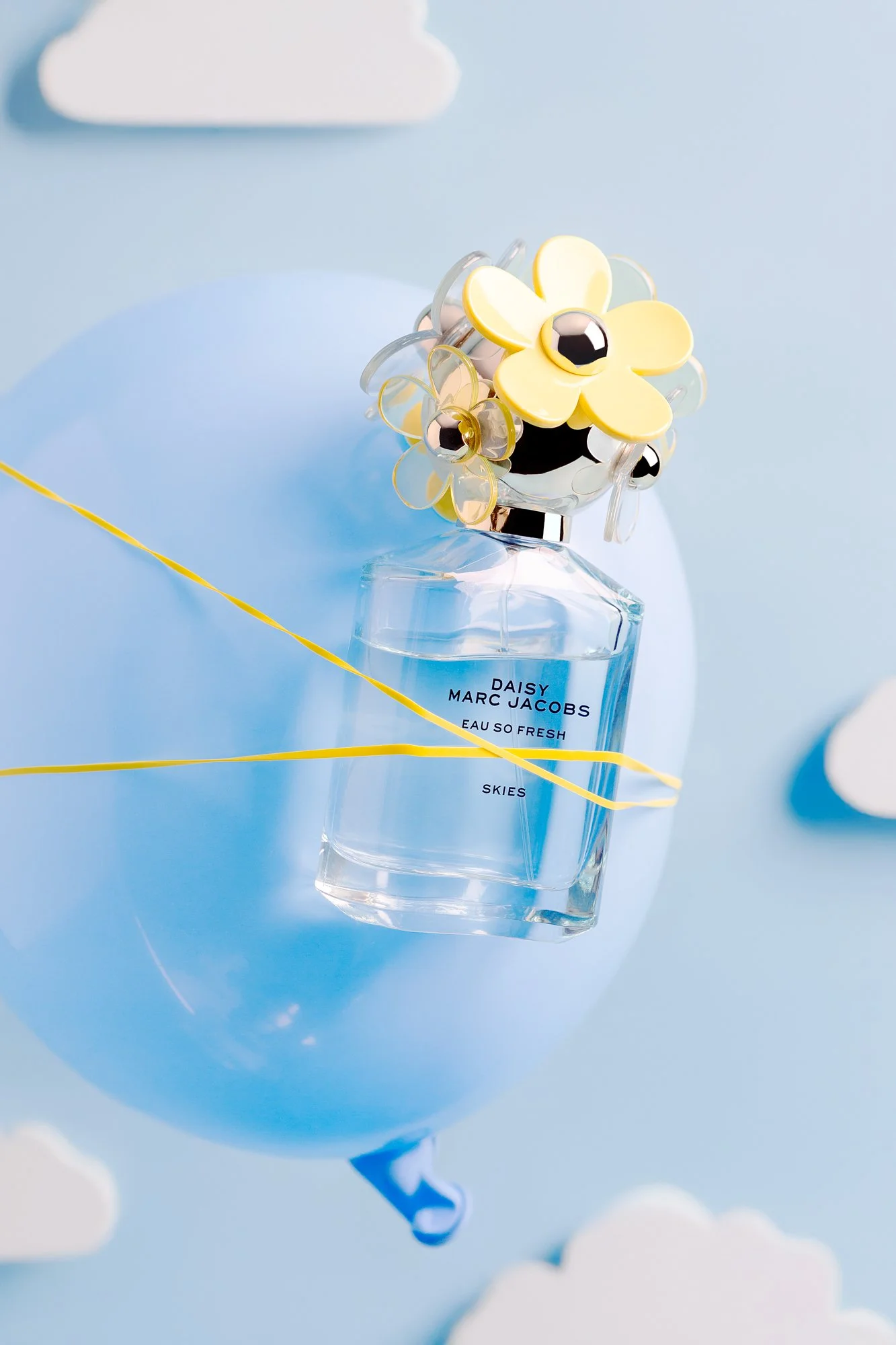 A Marc Jacobs Daisy Eau So Fresh perfume bottle with a flower-shaped cap, placed on a light blue balloon with clouds and string decorations in a sky-themed background.