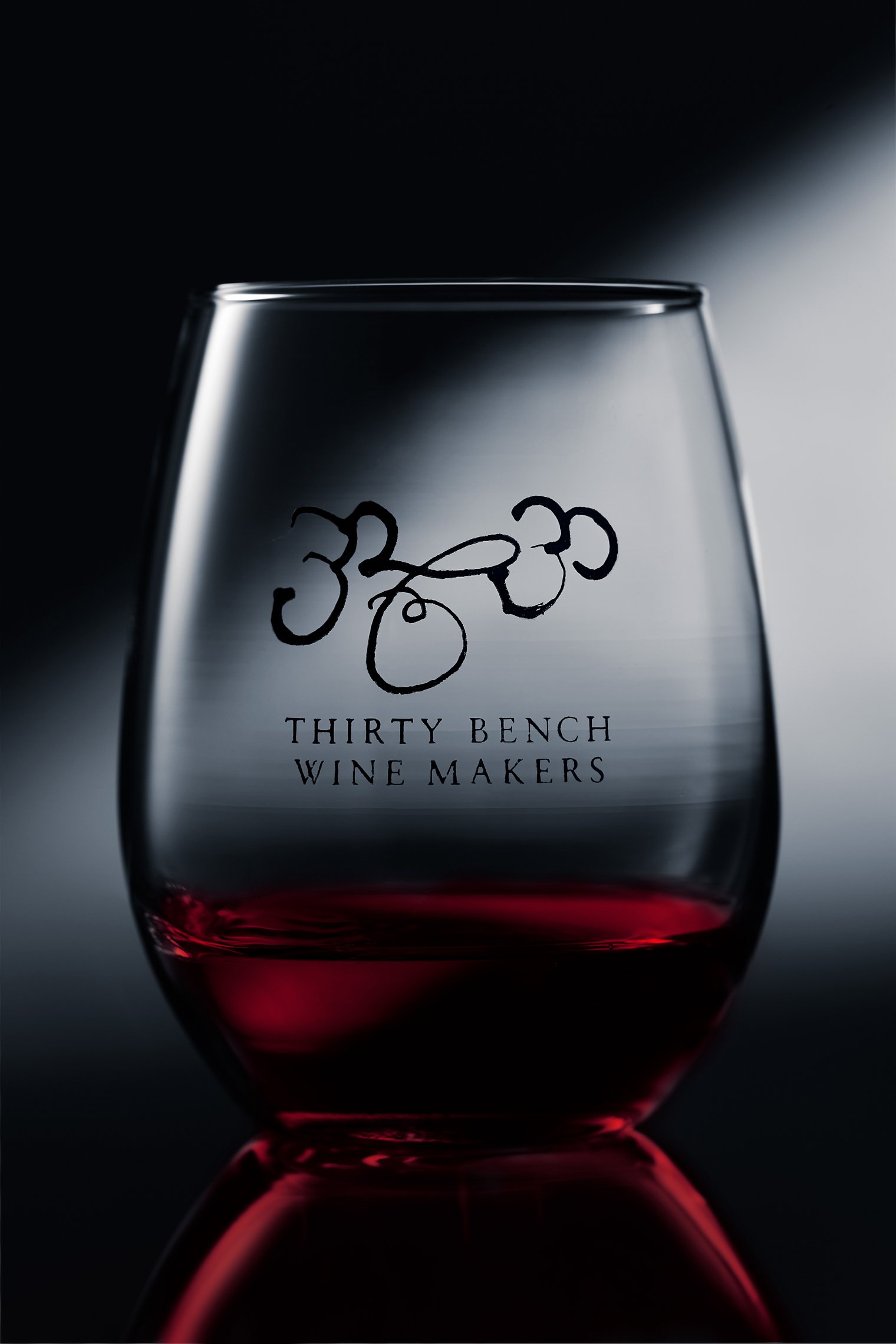 A clear wine glass with red wine at the bottom, inscribed with 'Thirty Bench Wine Makers' and abstract logo.