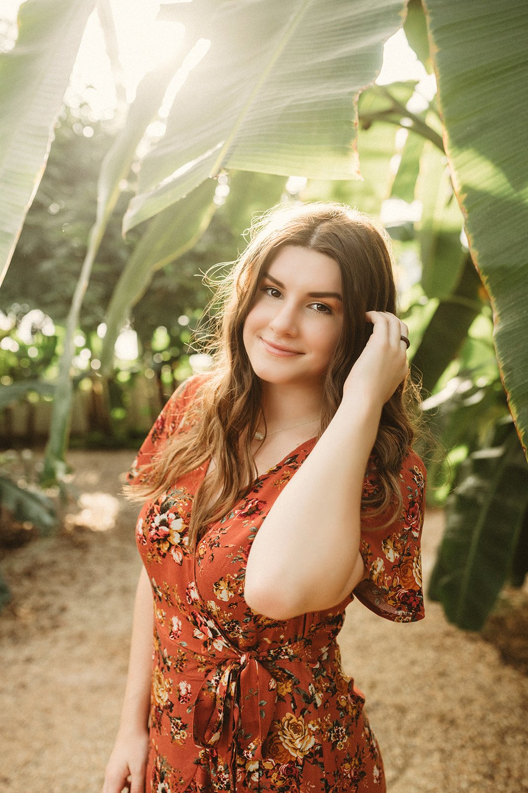 Woman in a floral dress standing among large green leaves, smiling in sunlight.
