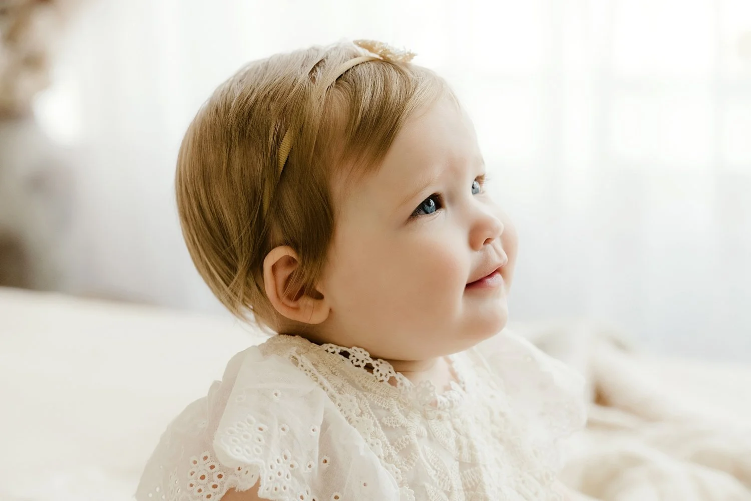 Profile of a baby with short blond hair wearing a white lace outfit.