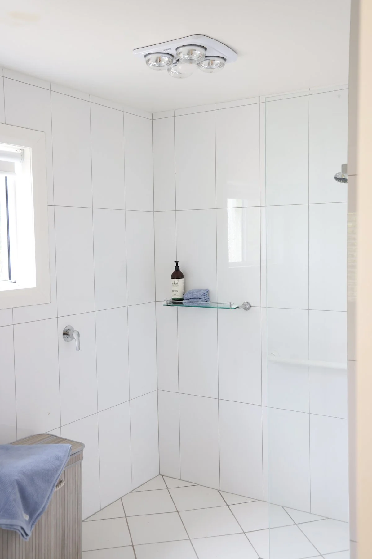 Clean white tiled shower area with a glass shelf holding a bottle of hand soap and blue towel, a small window, and a ceiling light fixture.