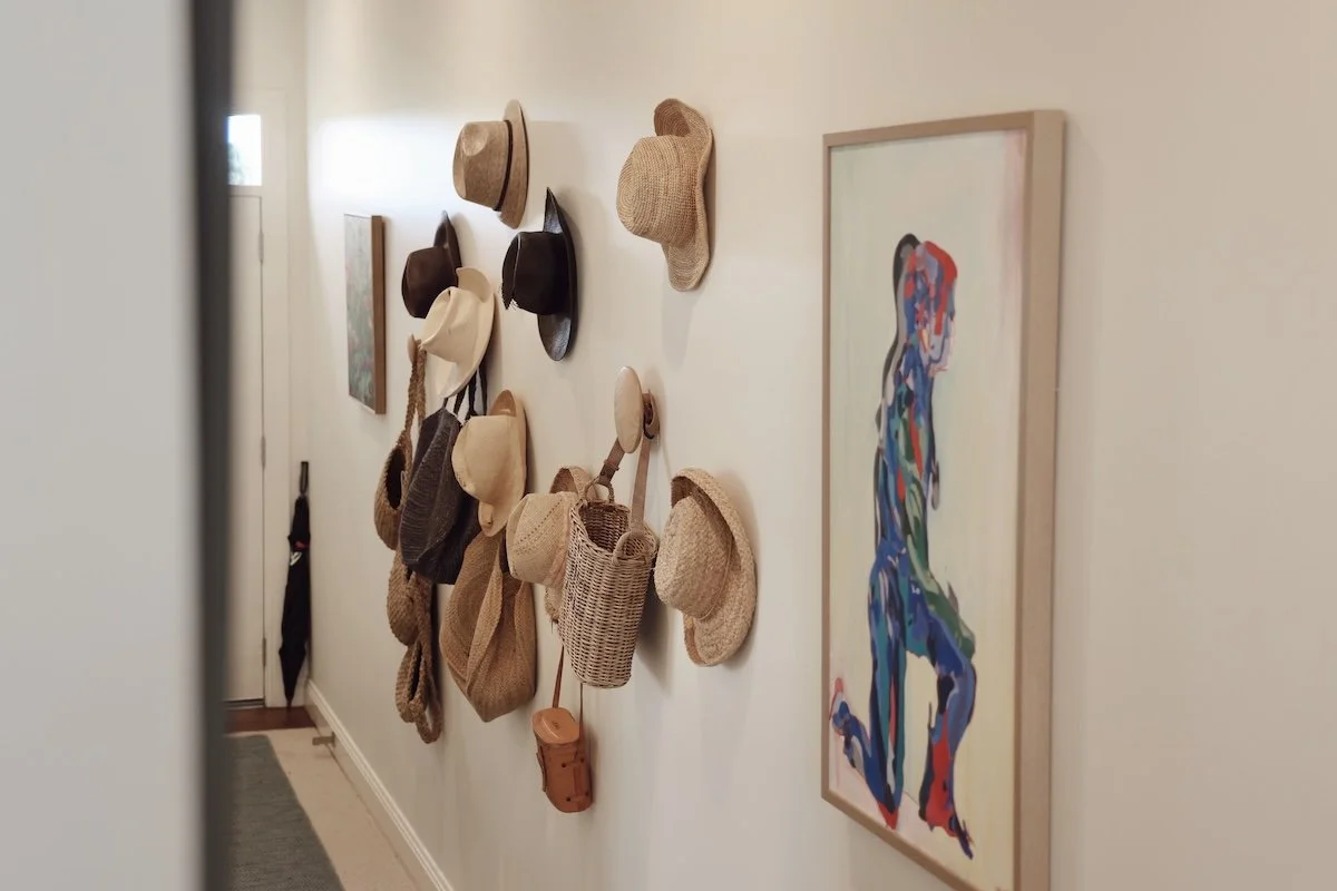 Hallway with a wall-mounted display of multiple woven and leather hats, and a colorful abstract painting of a woman.