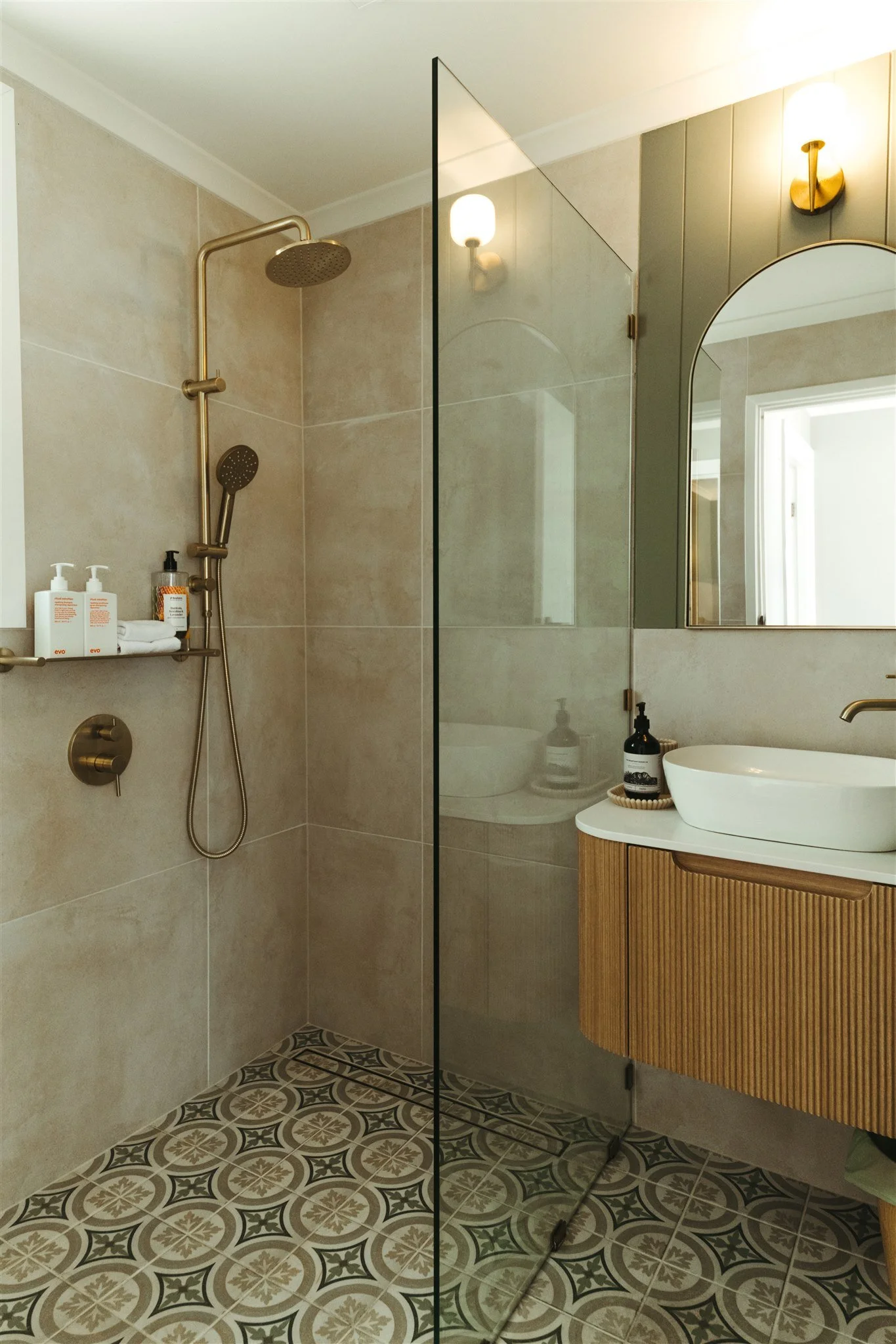 Modern bathroom with walk-in shower, beige tiled walls, decorative floor tiles, wooden vanity with vessel sink, large mirror, and wall-mounted lighting.