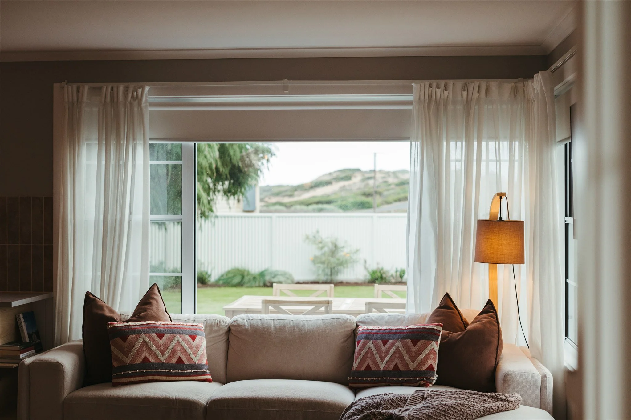 Living room view through open curtains showing a white sofa with patterned pillows, a floor lamp, and an outdoor patio with table and chairs against a grassy yard and hills in the background.