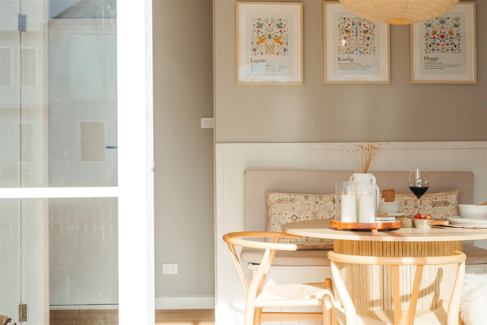 Dining area with a round wooden table, beige chairs, a bench with embroidered pillows, and framed artwork on the wall. The table has white candles, a vase with dried flowers, and a glass of red wine. Sunlight is coming through a glass door on the lef
