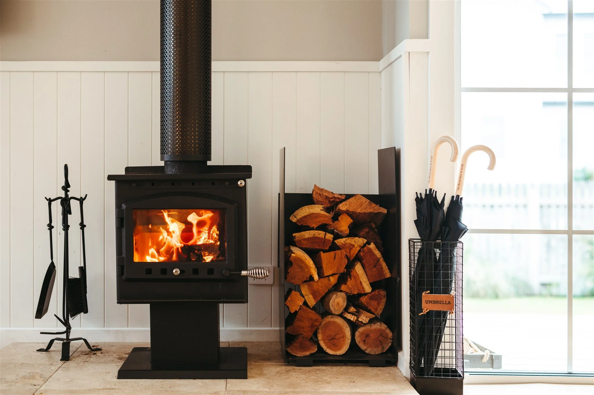 A black wood-burning stove with a fire inside, surrounded by a container of chopped firewood and a black wire umbrella stand holding black umbrellas and two white candy cane-style umbrellas by a sliding glass door.