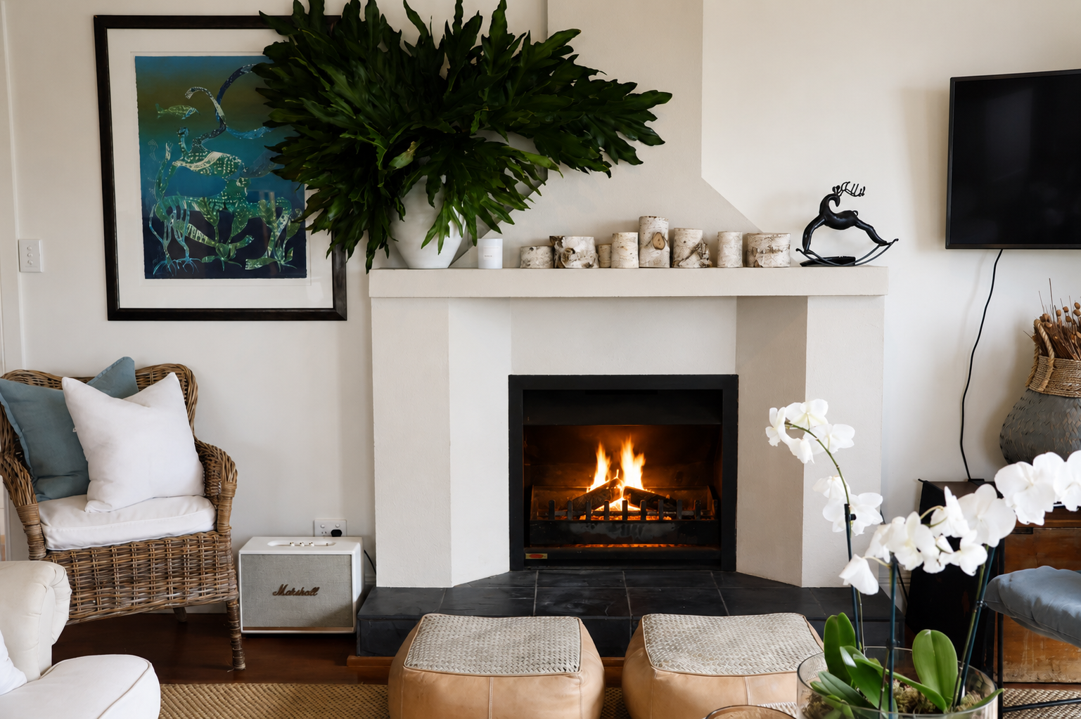 Living room with a fireplace, large green plant, framed colorful artwork, and decorative items including a black reindeer sculpture, white orchids, and a TV on the wall.