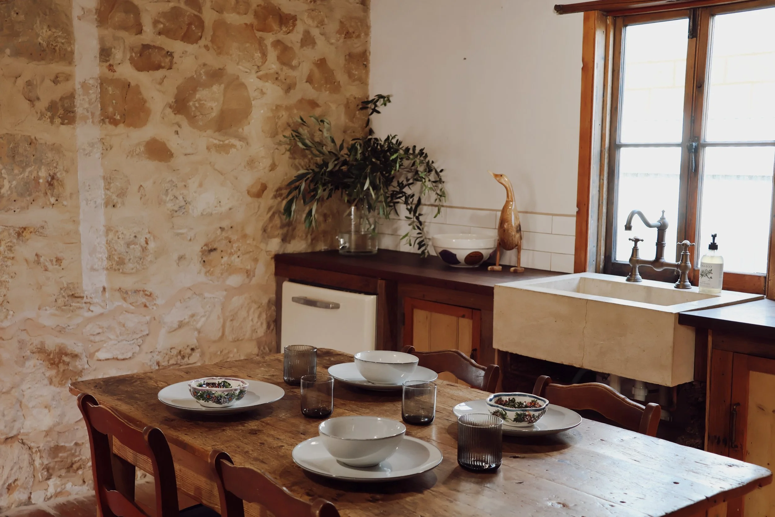 A rustic kitchen with a stone wall on one side, wooden cabinets, a white sink, a window with wooden frames, and a dining table set with bowls, glasses, and plates. There is a potted plant and decorative items on the countertop.