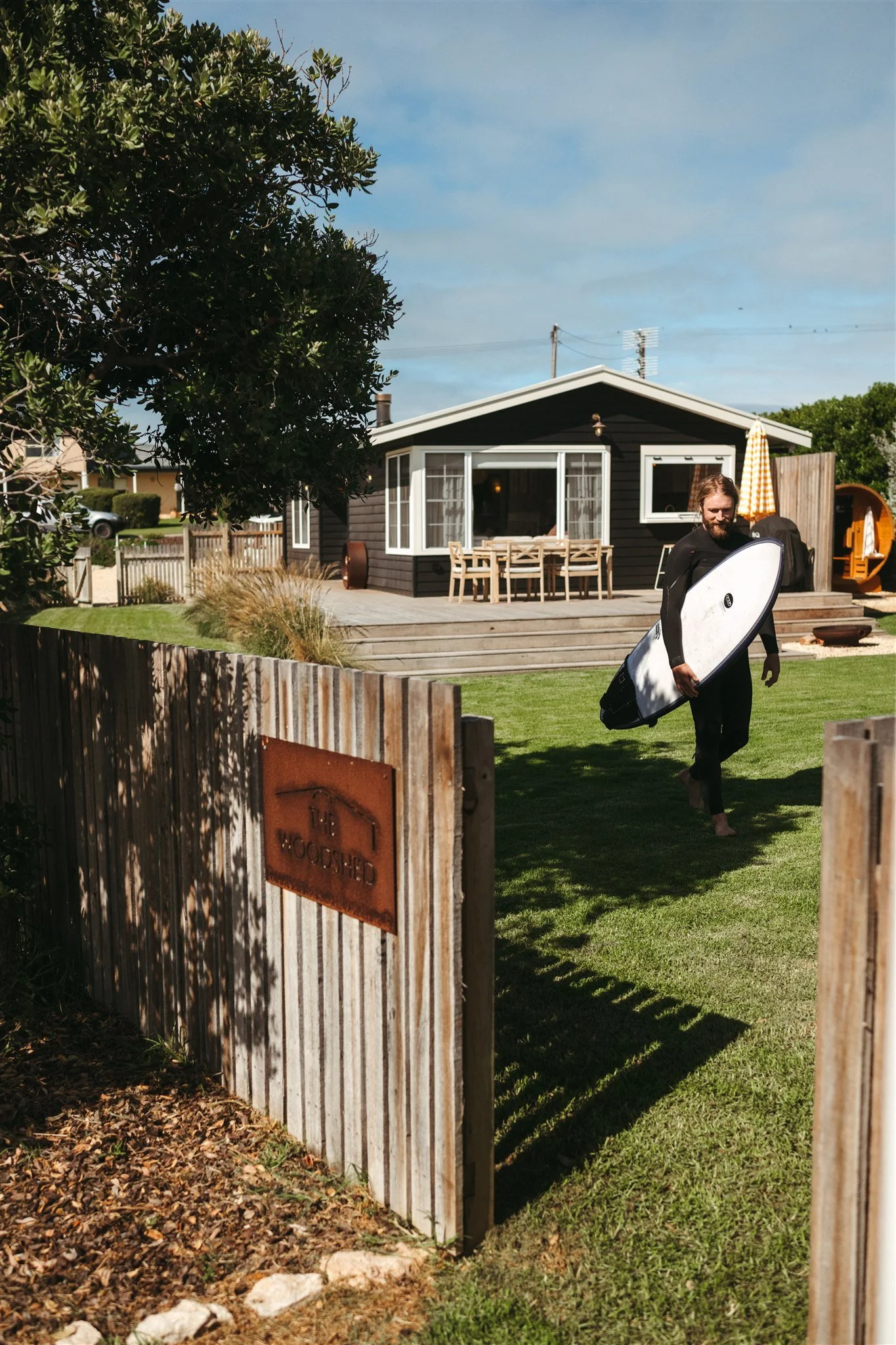 A man with a surfboard walking across a lawn towards a house with a deck, a wooden fence in front, and a sign that reads "The Wood Shed."