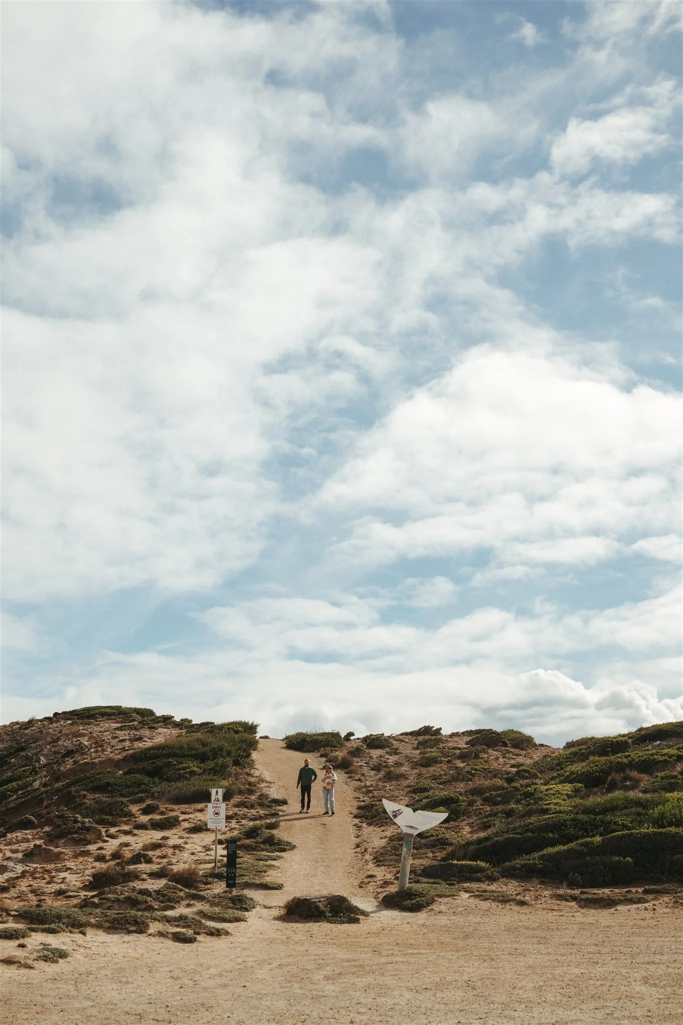 Two people walking up a dirt trail on a hill, surrounded by shrubbery, with a cloudy sky above.