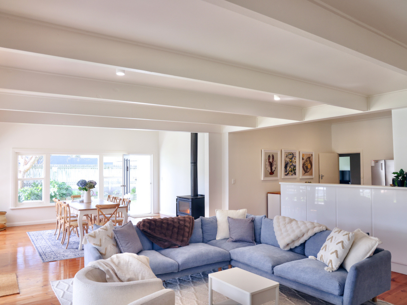 Bright living room with a blue sectional sofa, white armchair, wooden dining table with chairs, artwork on the wall, and a wood stove.