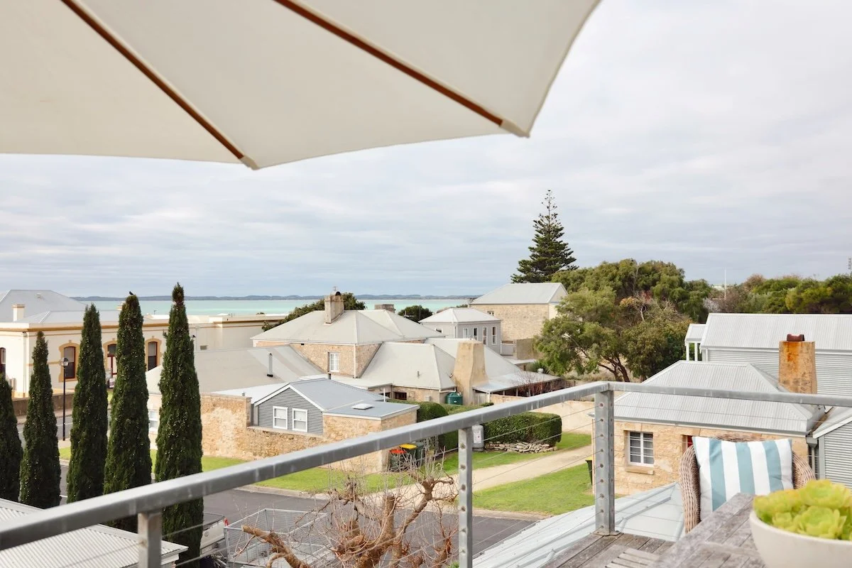 View from a balcony showing nearby houses with grey roofs, a street, a row of tall cypress trees, and a distant body of water under a cloudy sky, with an umbrella, a chair with a striped pillow, and a small table with a green plant in the foreground.