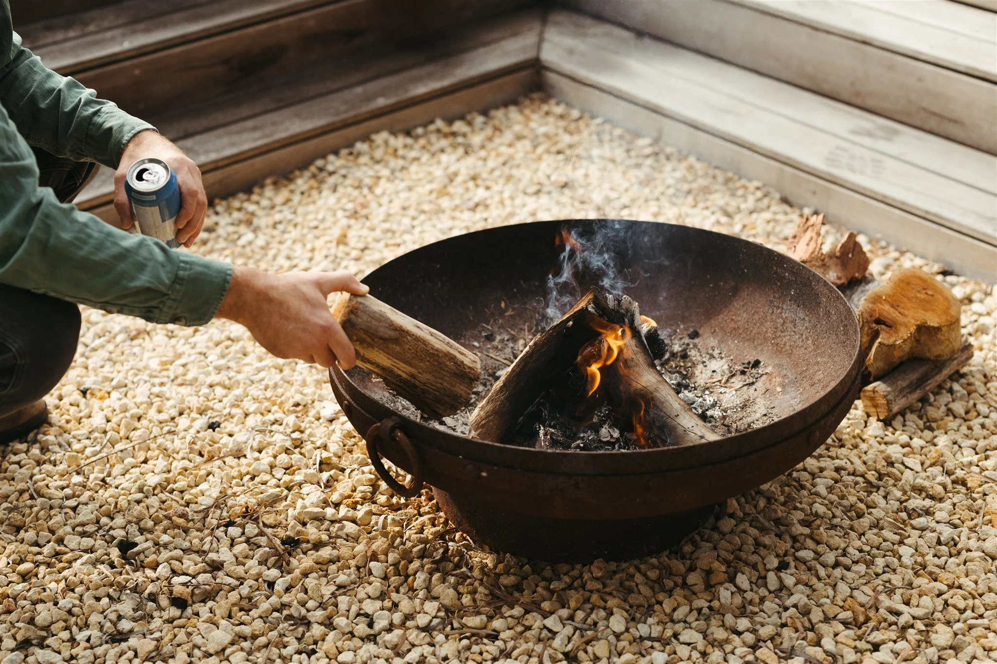 Person tending a small campfire with wood logs inside a black fire pit on gravel ground, with one log in hand and a soda can nearby.
