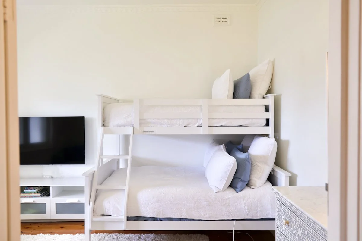 A bedroom with a white bunk bed and white bedding, with pillows on both beds, placed in front of a flat-screen TV and white entertainment unit.