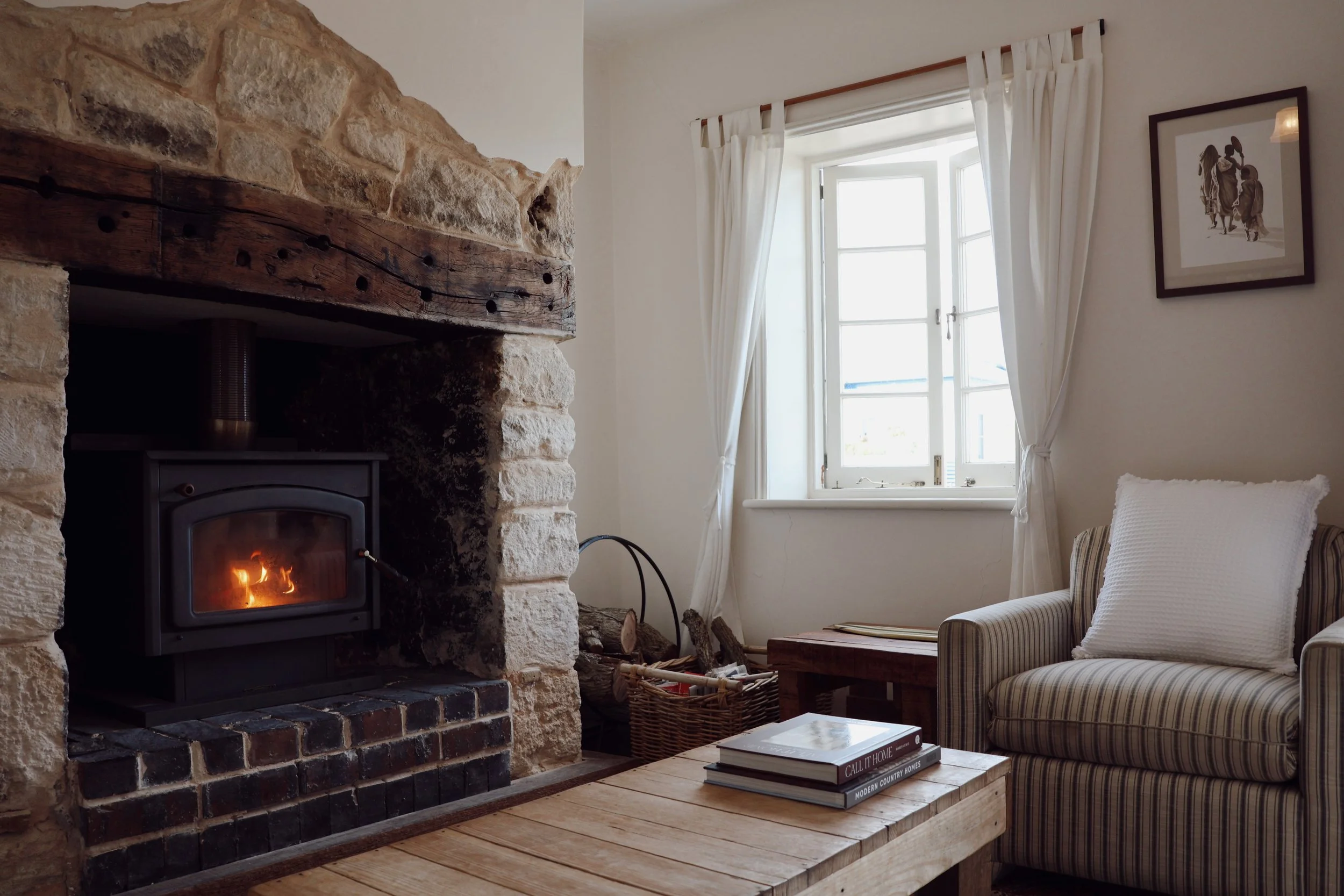 Living room with a lit wood stove, stone walls, a wooden mantle, a window with white curtains, a striped armchair with pillows, a basket of firewood, and a coffee table with books.