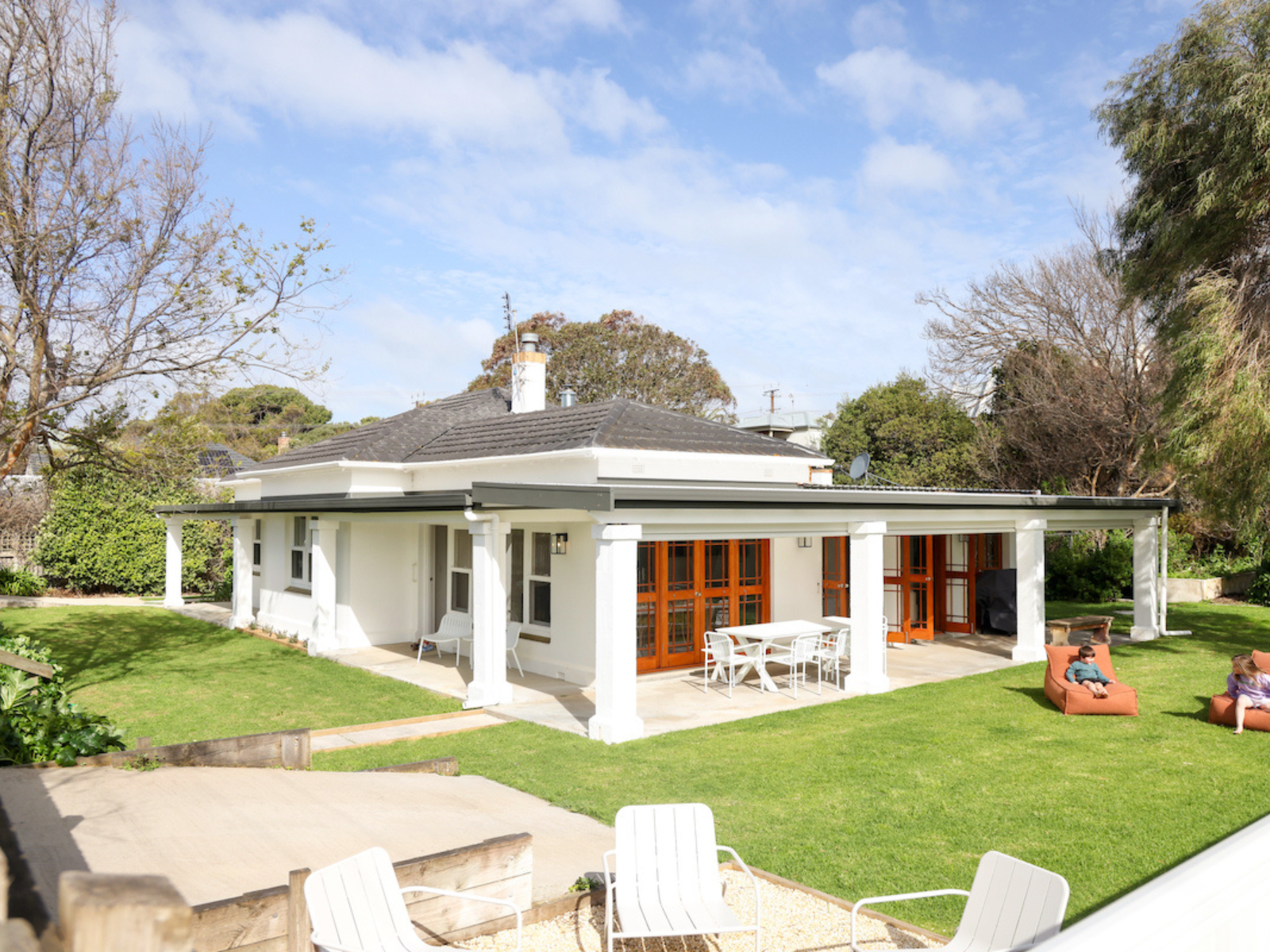 A backyard view of a white house with a covered patio area, surrounded by green grass and trees, with two children playing on outdoor furniture.