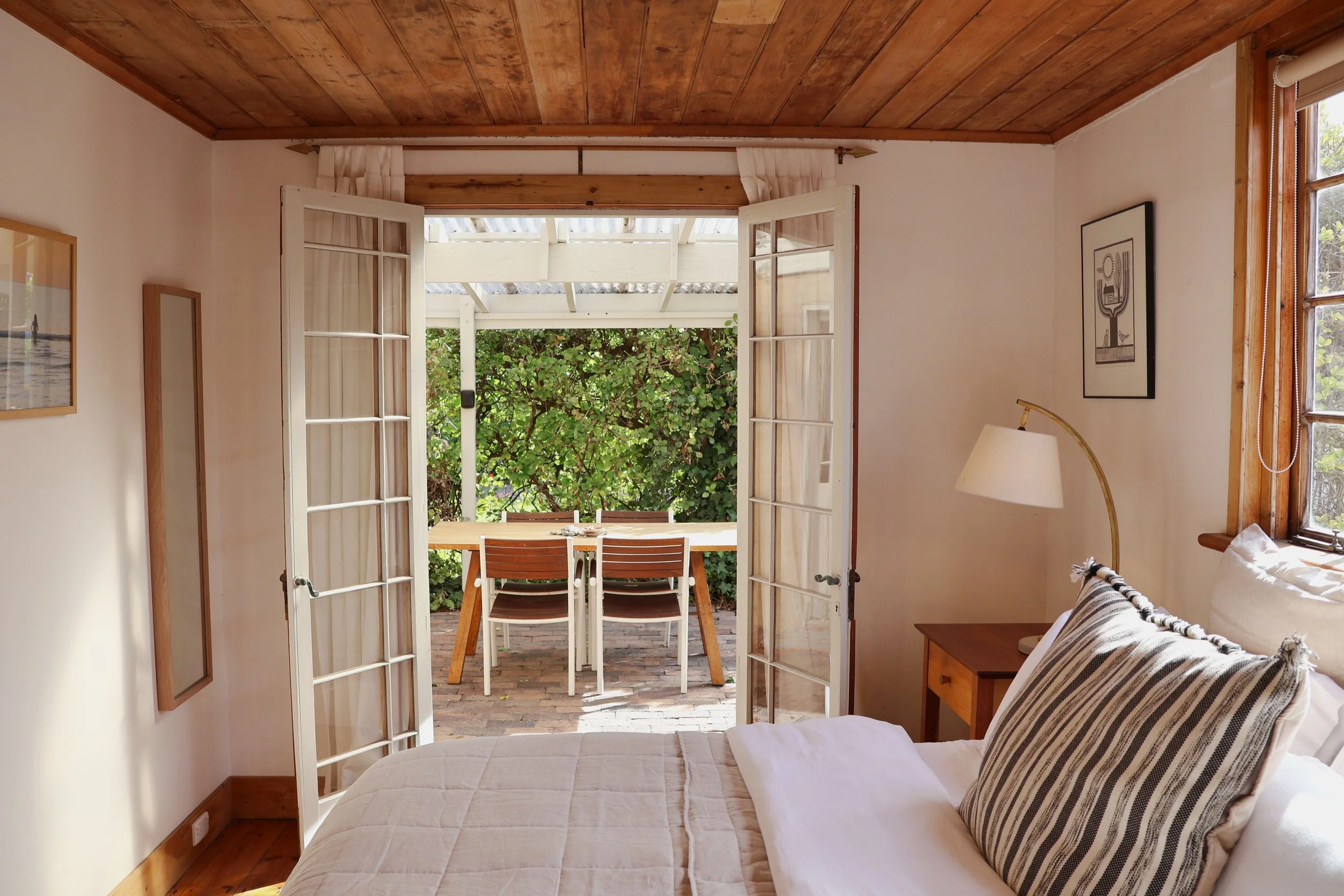 View from inside a bedroom looking out a set of open French doors to a patio with a wooden dining table and chairs, surrounded by green bushes and trees.