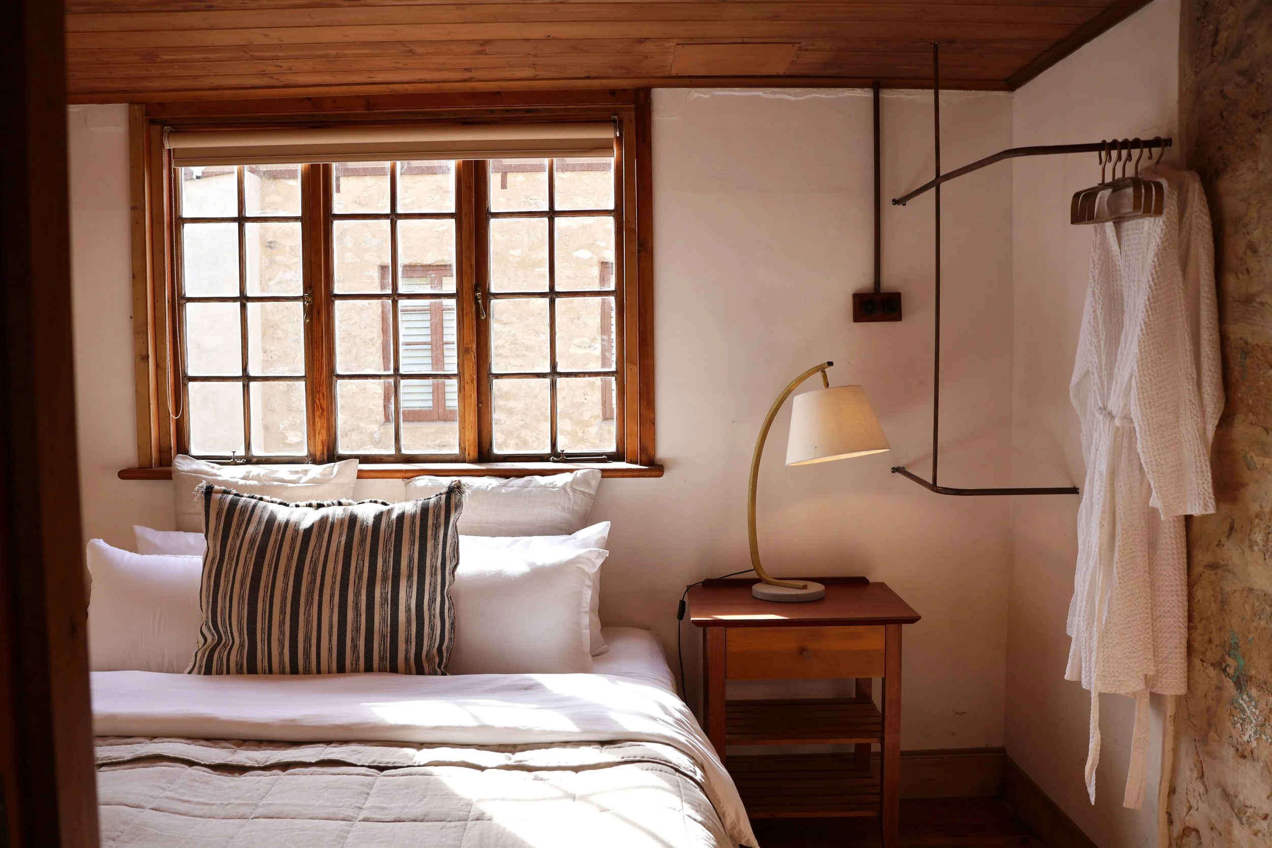 Interior view of a cozy bedroom featuring a bed with white linens and striped pillows, a wooden bedside table with a beige lamp, a large window with wooden framing allowing natural light, a beige wall with a mounted black metal shelf with hangers, an