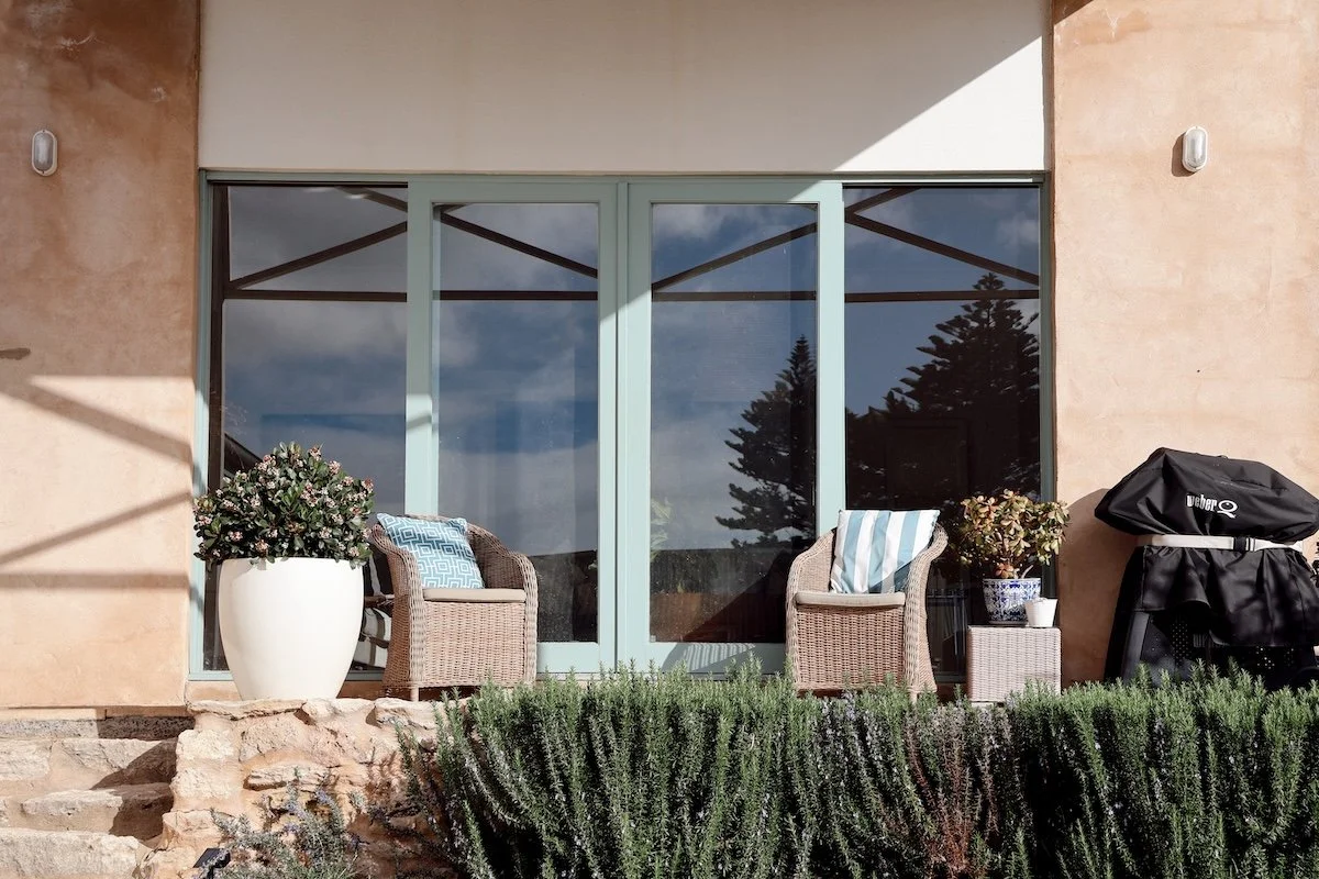 Outdoor patio with two wicker chairs and colorful cushions, potted plants, and a barbecue grill on the right, in front of a large glass sliding door reflecting trees and sky.