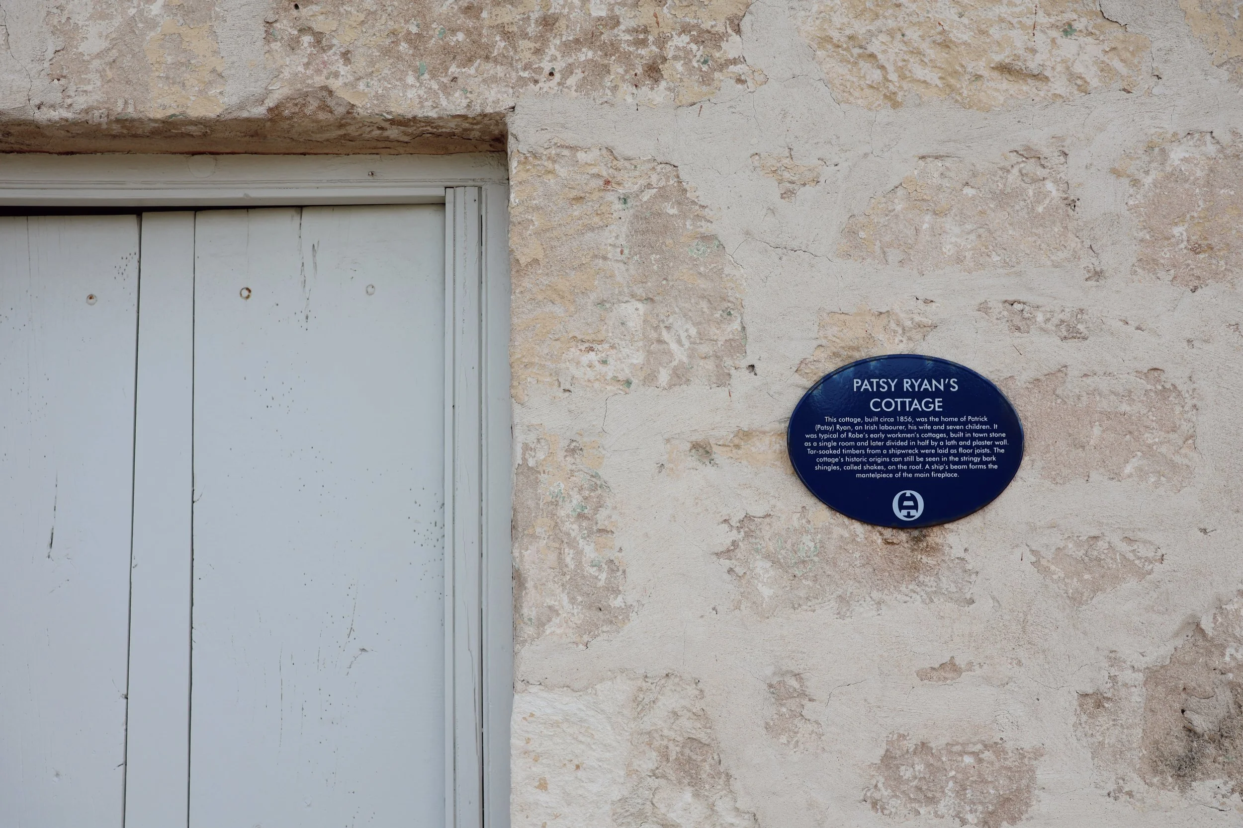 Photo of a textured, weathered beige stone wall with a white door on the left and a blue informational plaque on the right. The plaque provides historical information about Patsy Ryan's Cottage, built around 1856.