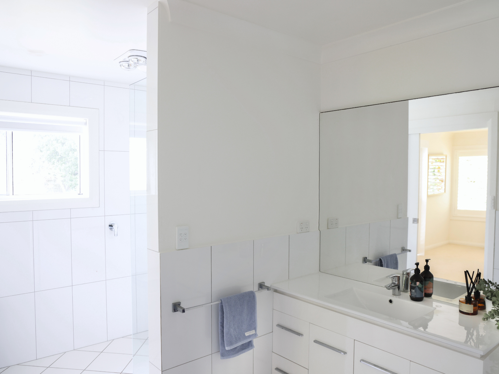 A modern white bathroom with a large mirror, sink, and toiletries, with a window providing natural light.