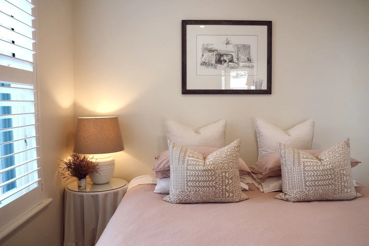 A cozy bedroom with a bed featuring multiple pillows, a bedside table with a lamp, and framed artwork on the wall.