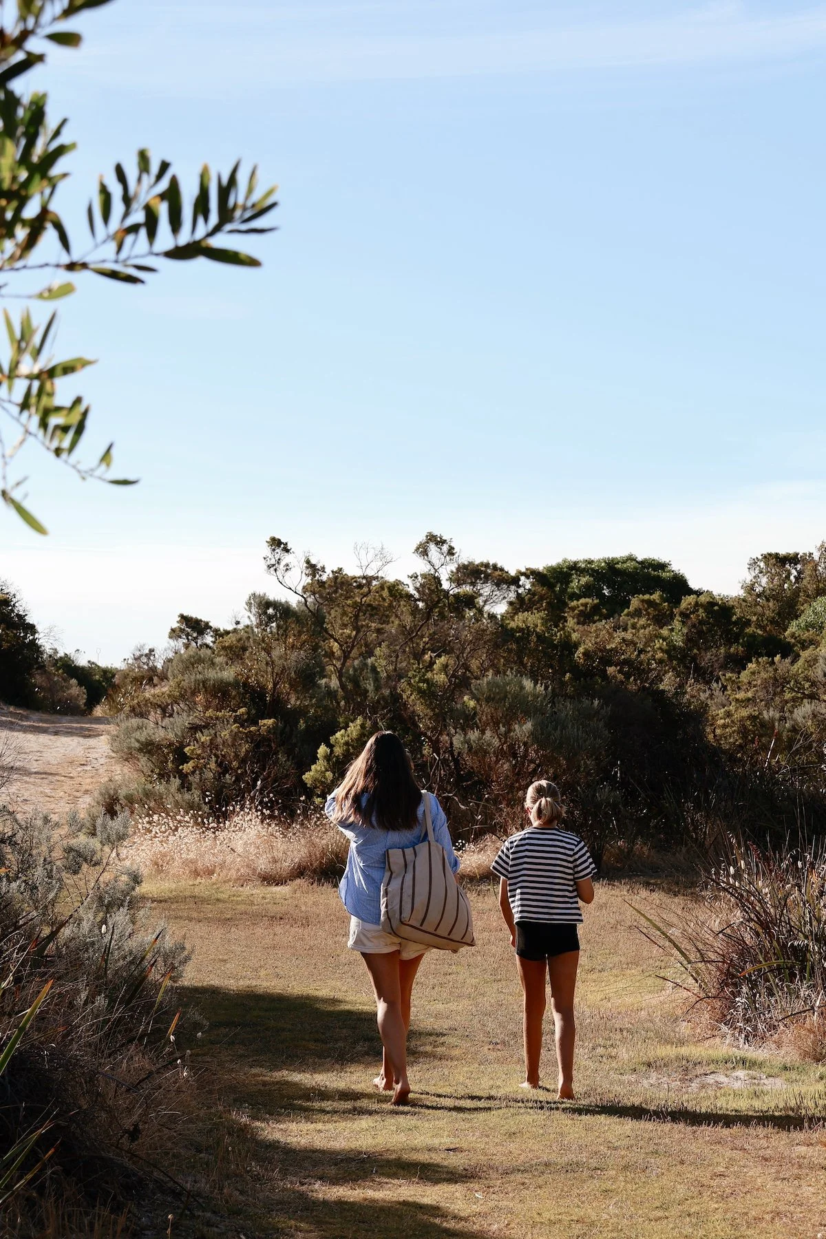Two women walking barefoot on a dirt trail in a grassy and bushy landscape, under a clear blue sky.