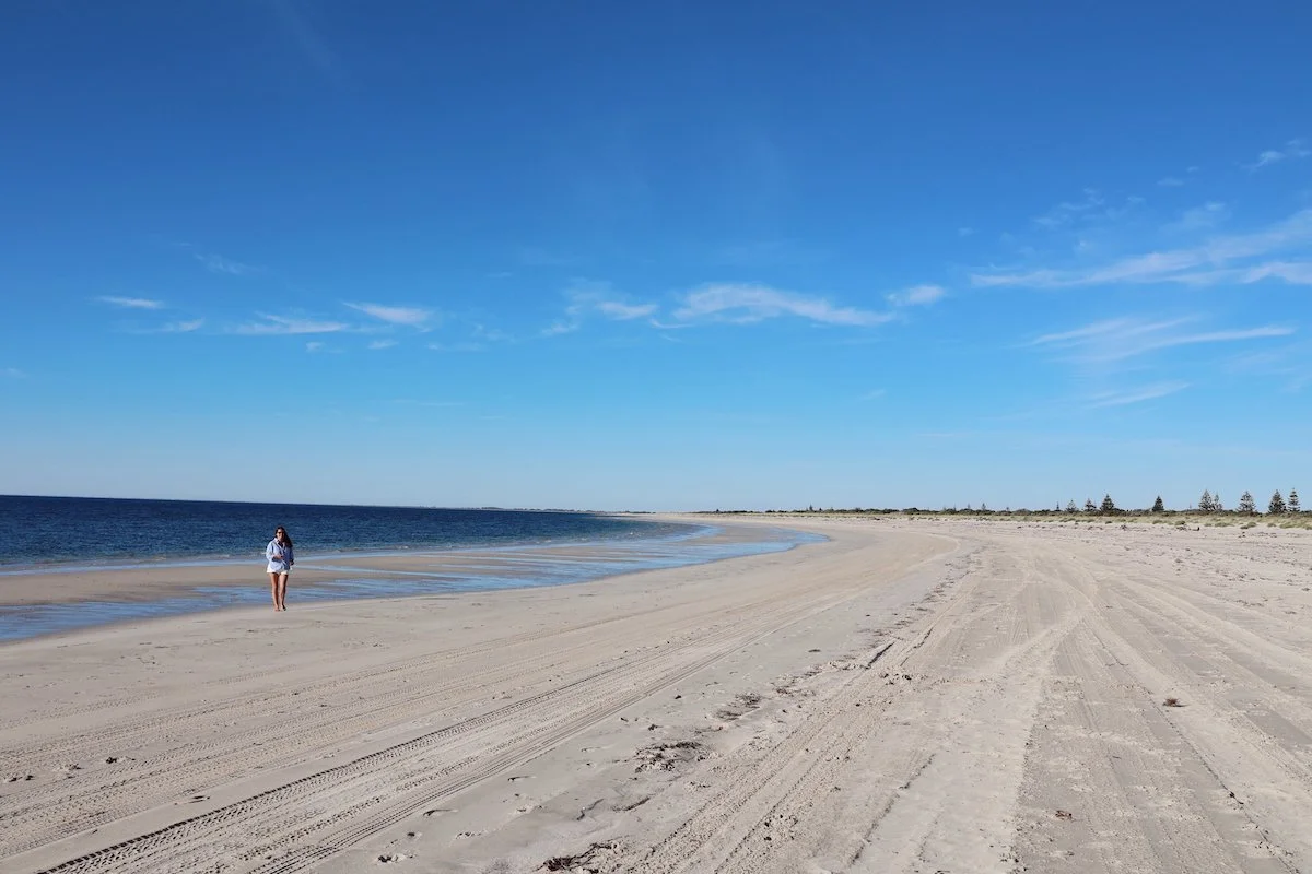 A woman walking alone on a wide, empty sandy beach under a bright blue sky with a few clouds, with the ocean on one side and distant trees on the horizon.
