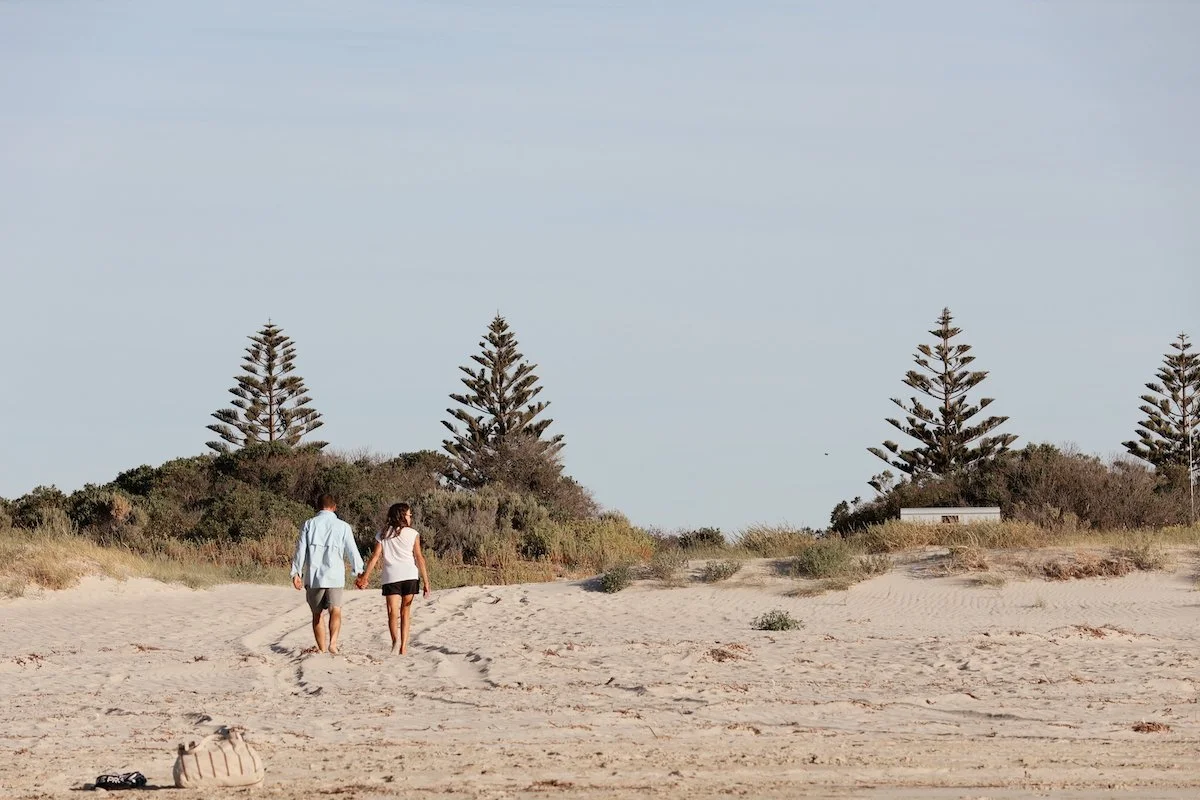 A couple walking hand in hand on a sandy beach with grasses and pine trees in the background.