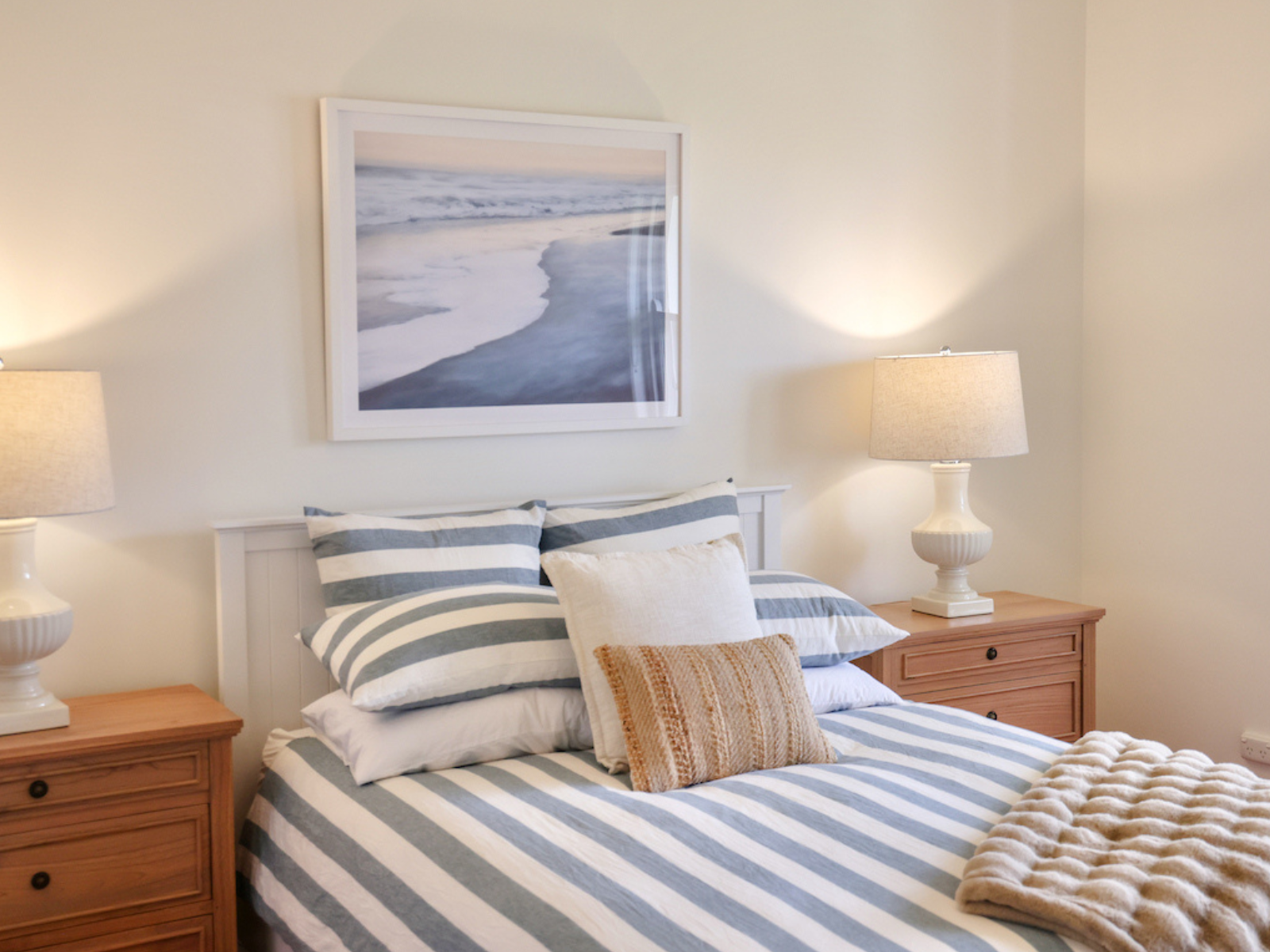 A cozy bedroom with a bed covered in blue and white striped bedding, topped with multiple pillows, against a light-colored wall. Two wooden nightstands with beige lamps are on either side of the bed. A framed ocean scene hangs above the headboard.