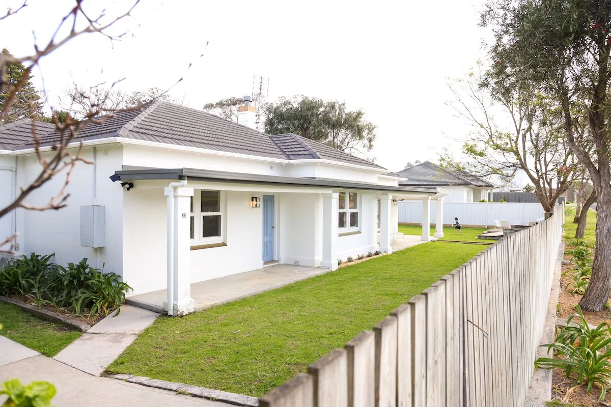 White house with blue door, gray roof, and a fenced backyard with green grass and trees.