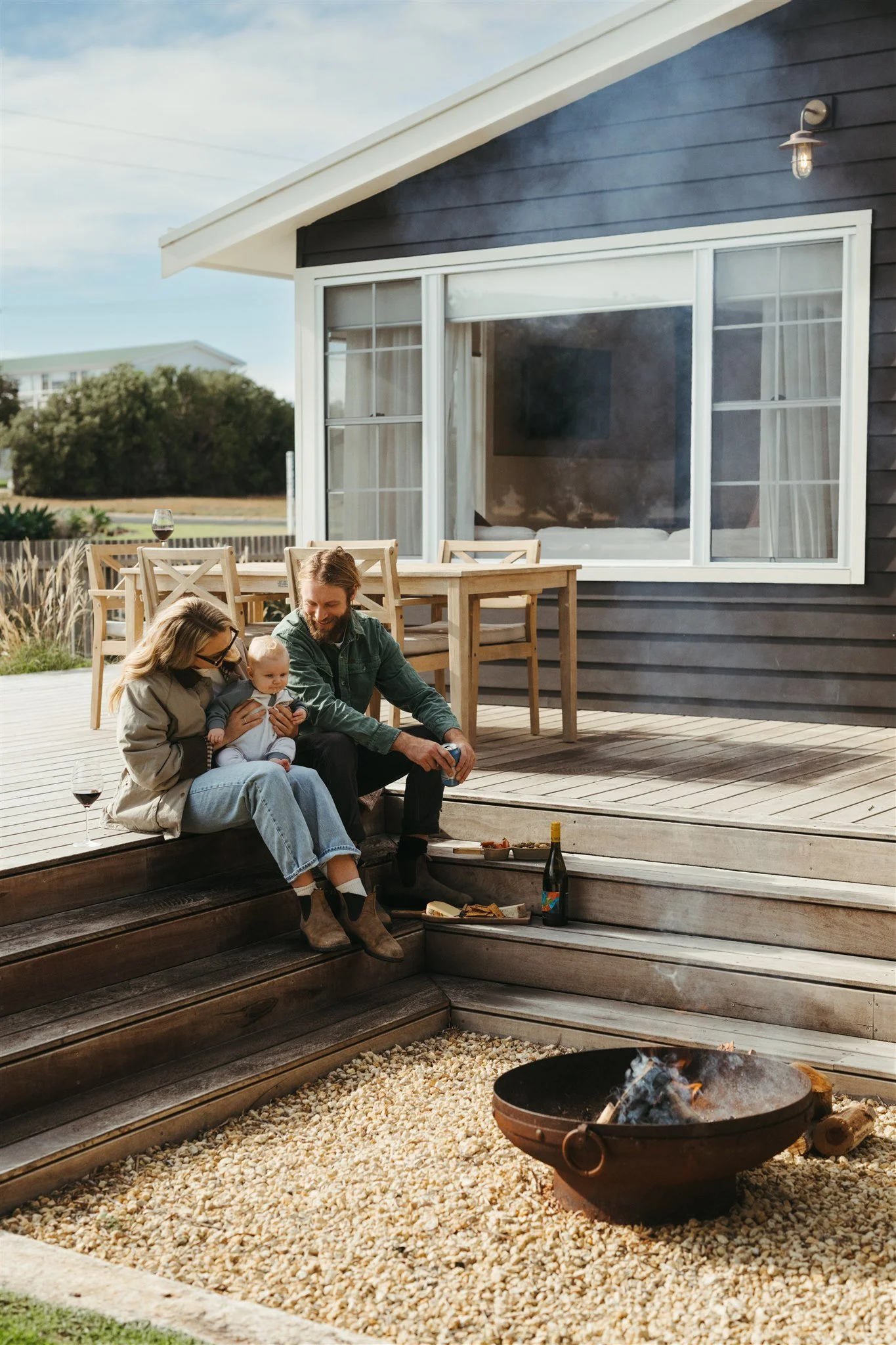 Family sitting on wooden steps outside a house, enjoying a bonfire with a bottle of wine and snacks.