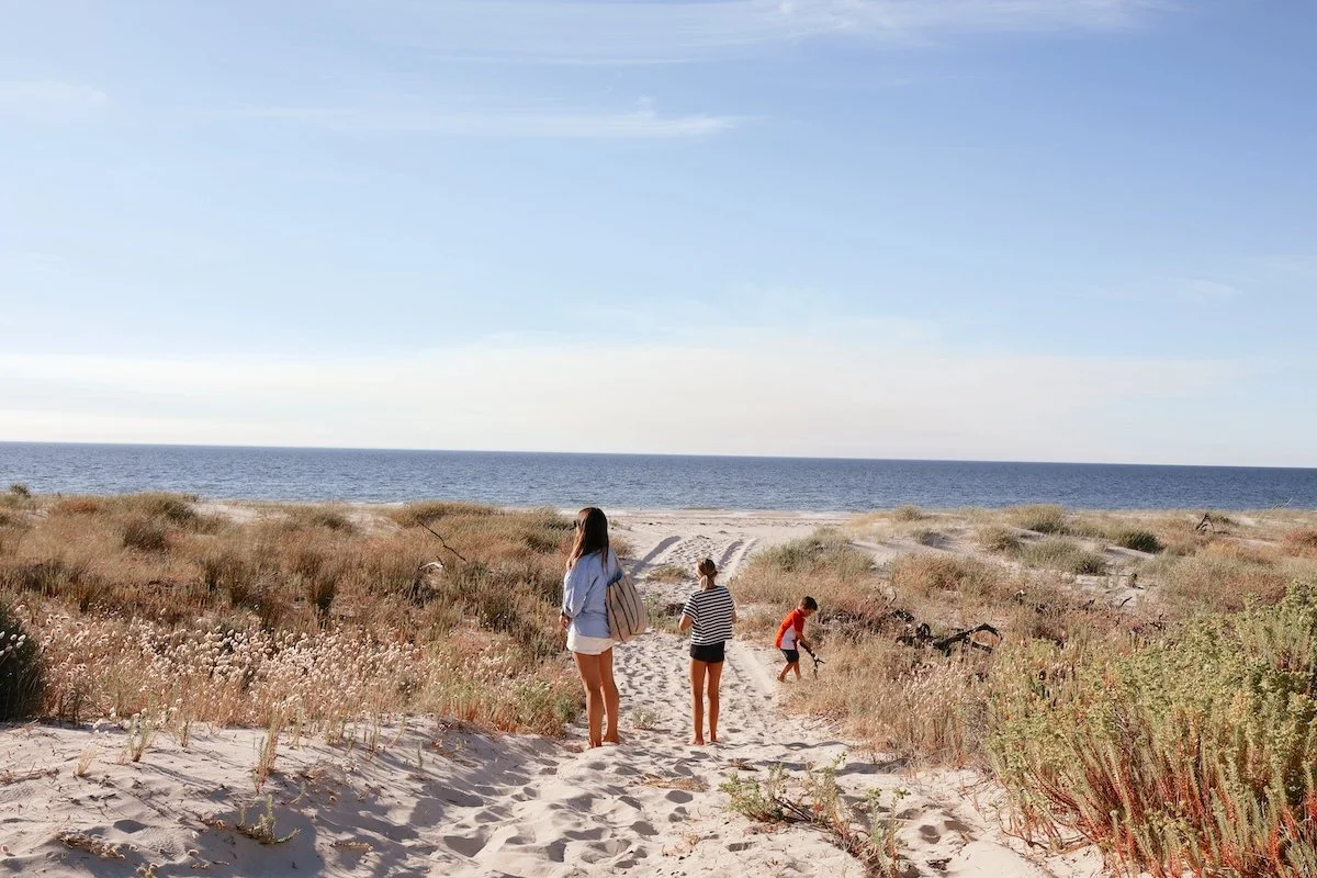 People walking on sandy beach dunes toward the ocean under a partly cloudy sky