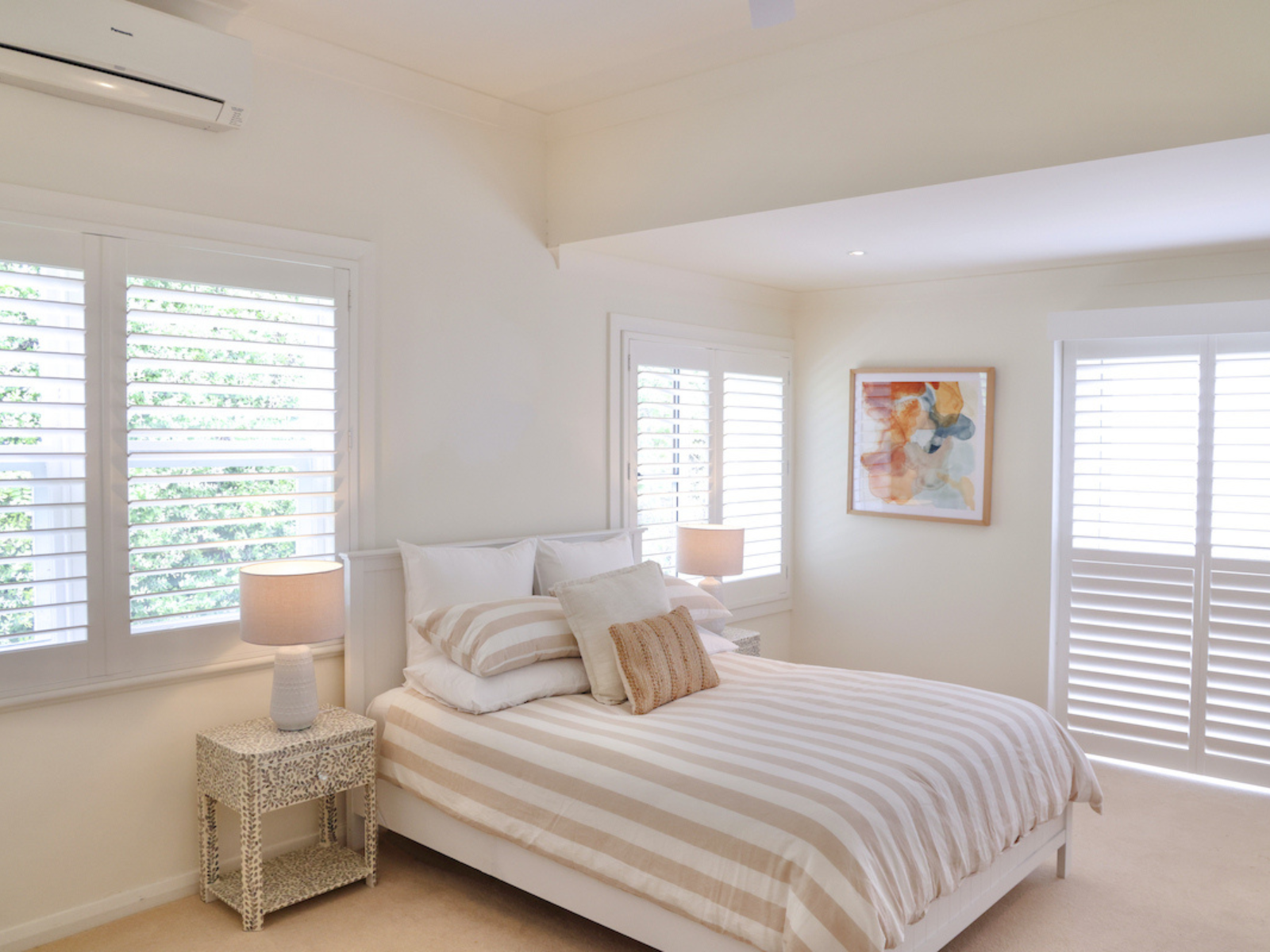 A bedroom with a white bed with beige-striped bedding, a nightstand with a white lamp, and abstract artwork on the wall. Multiple windows with white blinds allow natural light.