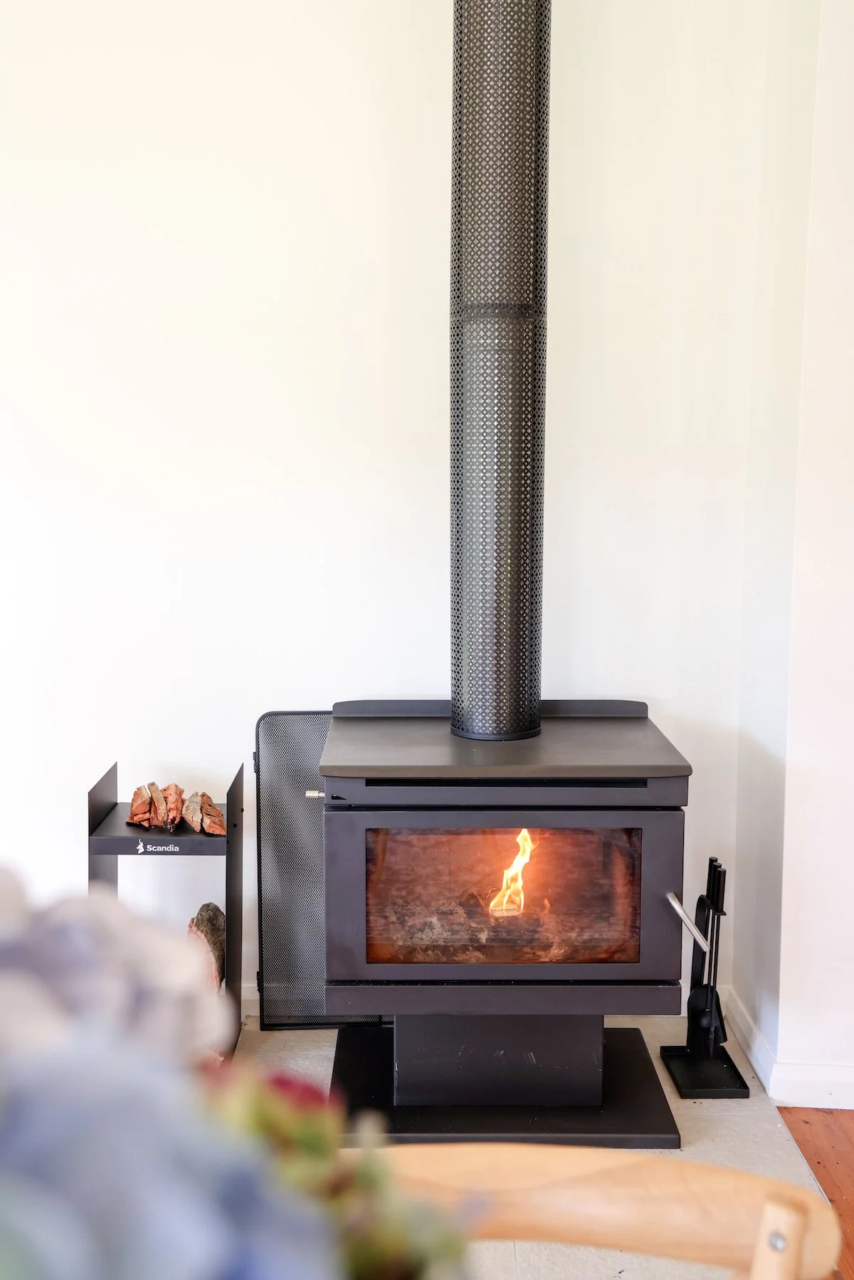 A modern living room with a black fireplace showing a fire inside, a black metal chimney pipe above, and a small wood log holder with wood on the left side, against white walls.