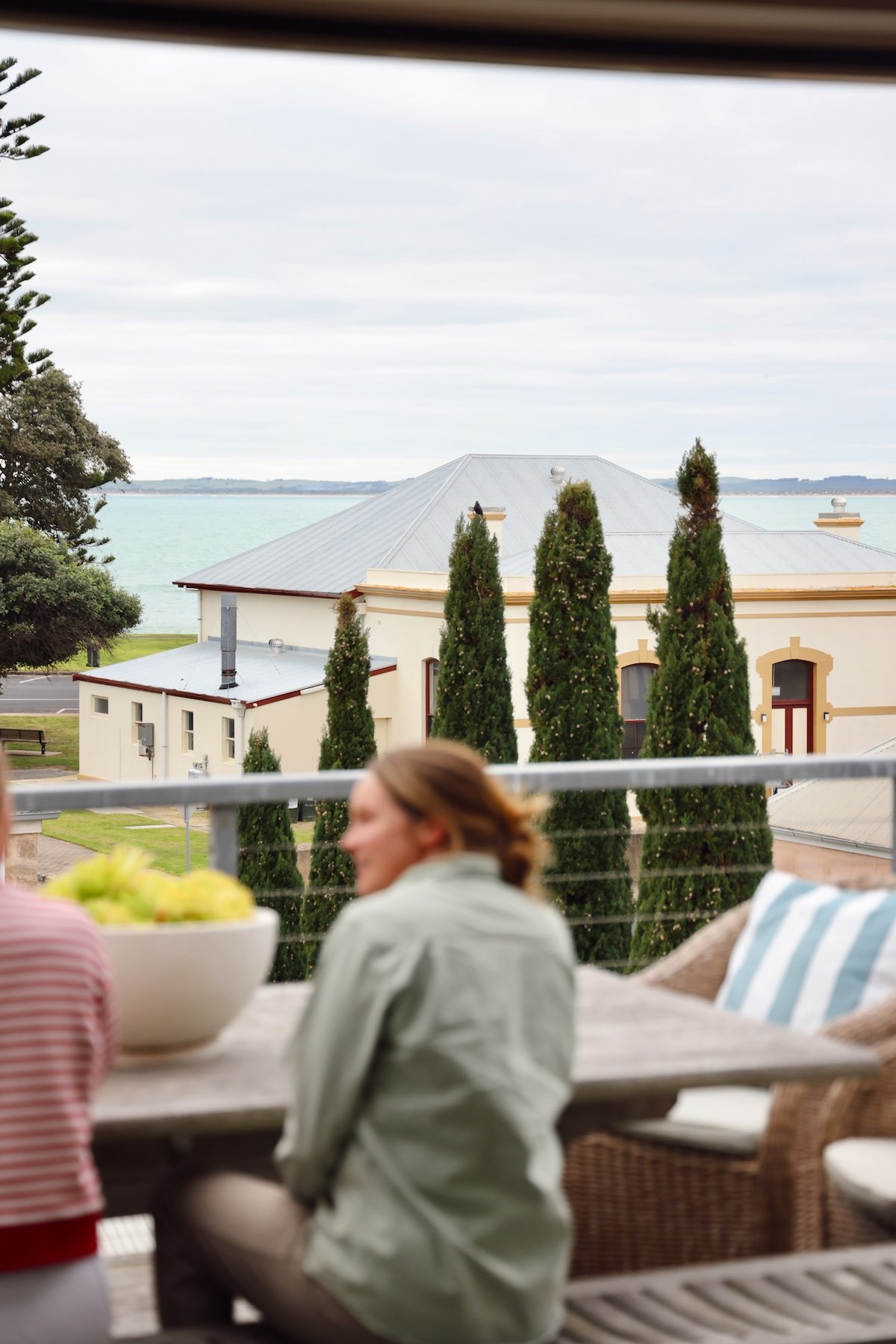People sitting at a table on a balcony, with a view of a house with a gray roof, tall trees, and a body of water in the background under a cloudy sky.