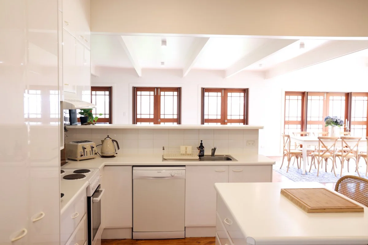 Bright kitchen with white cabinets and countertop, view toward dining area with wooden table and chairs, multiple wooden framed windows bringing in natural light, potted plant near windows.