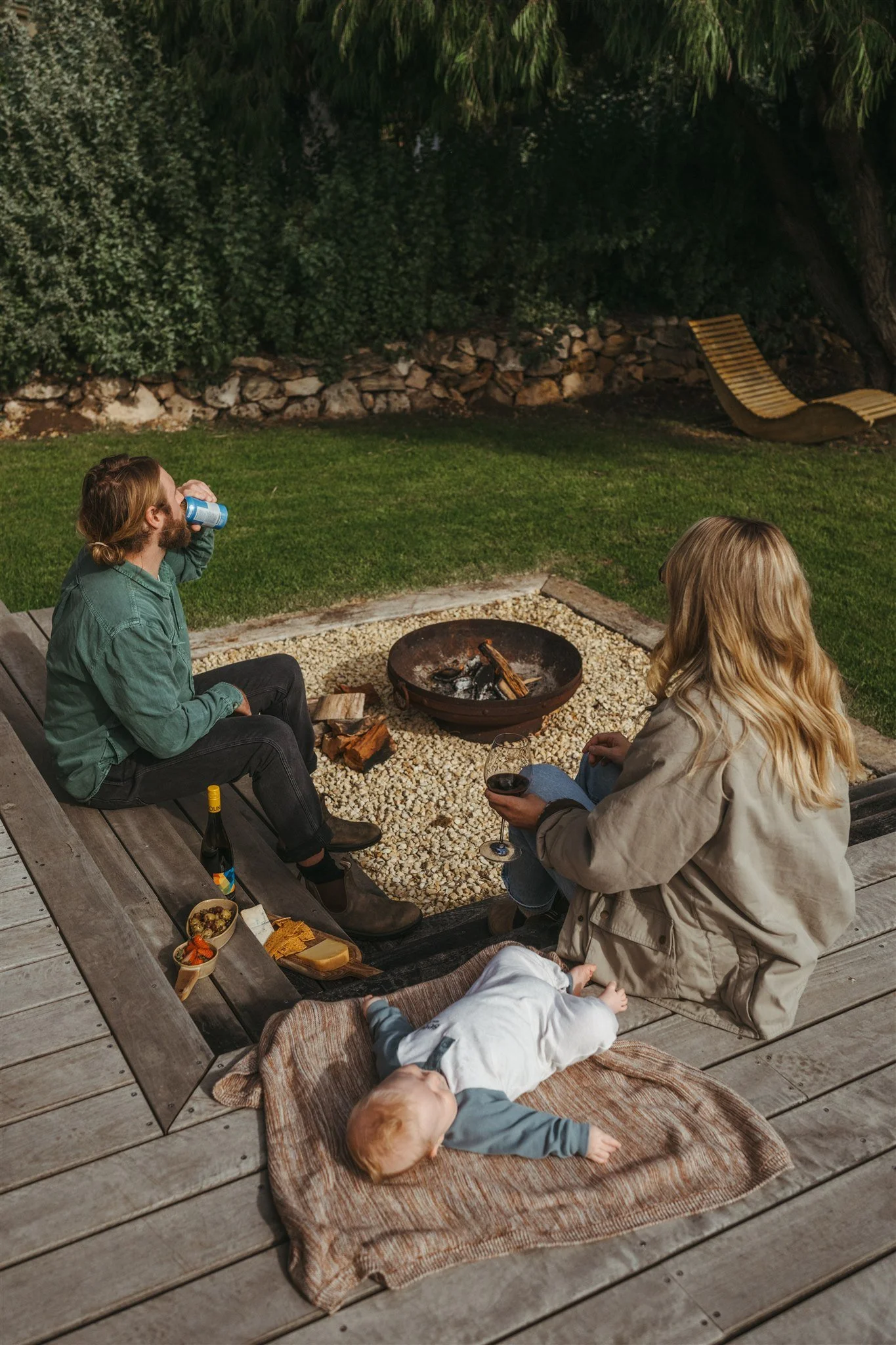 A family gathered around a backyard fire pit at night, with one person drinking from a can, another holding a glass of wine, and a baby lying on a blanket nearby. Snacks and a bottle are on the wooden deck.
