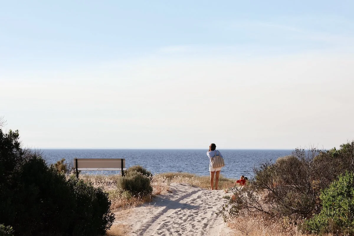 A woman standing on a sandy path overlooking the ocean with a bench nearby and shrubs on either side of the path, a child sitting in the sand to her right.