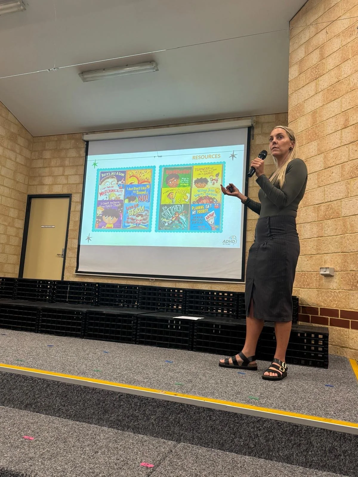 Eliza Dadson presenting to eductors about ADHD, the current slide is titled 'Resources'. She is holding a microphone and a remote control, standing on a small stage.