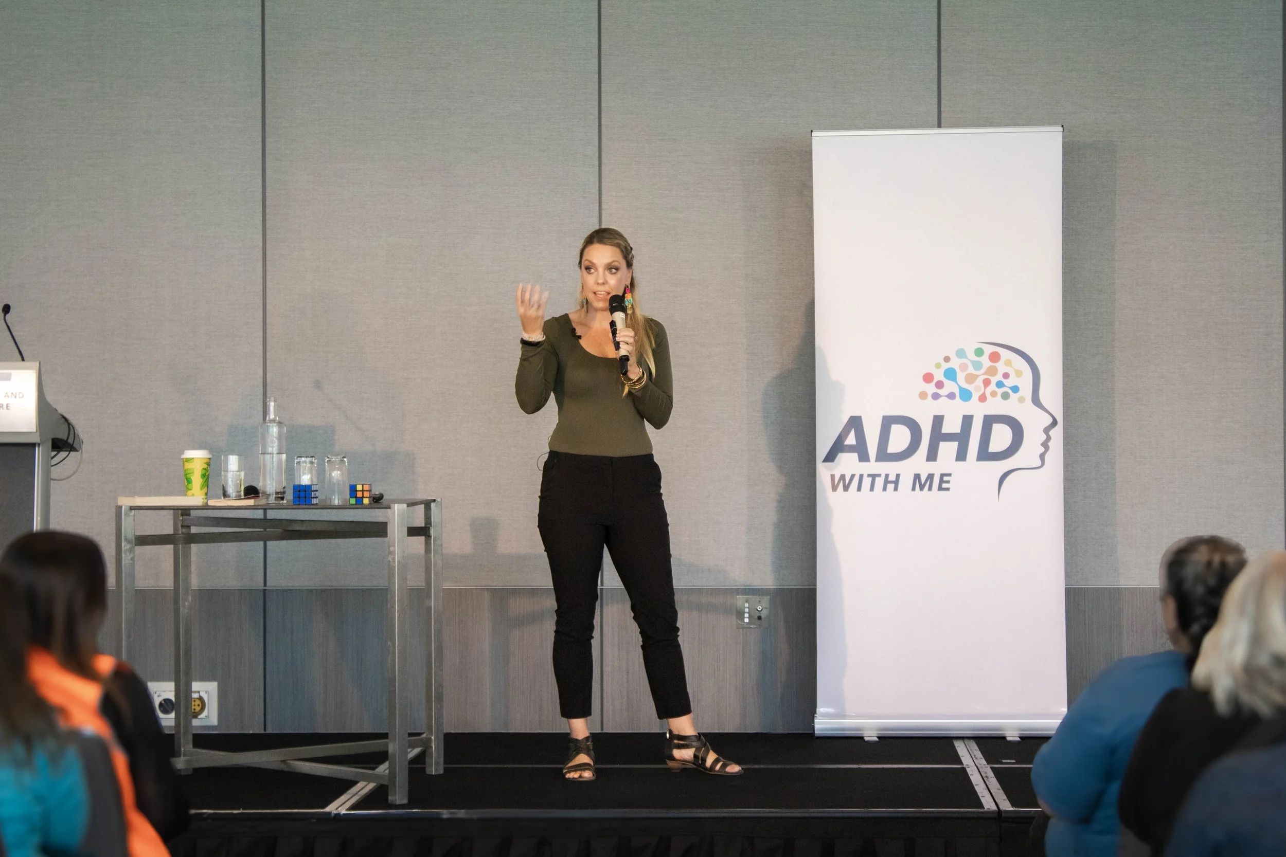 A woman gives a presentation at a conference, standing on a stage with a microphone in her right hand and gesturing with her left hand, next to a banner that reads 'ADHD WITH ME' with a stylized profile and colorful brain illustration. She is wearing a green top, black pants, and black sandals, and there is a table with jars, water bottles, and a Rubik's Cube nearby.