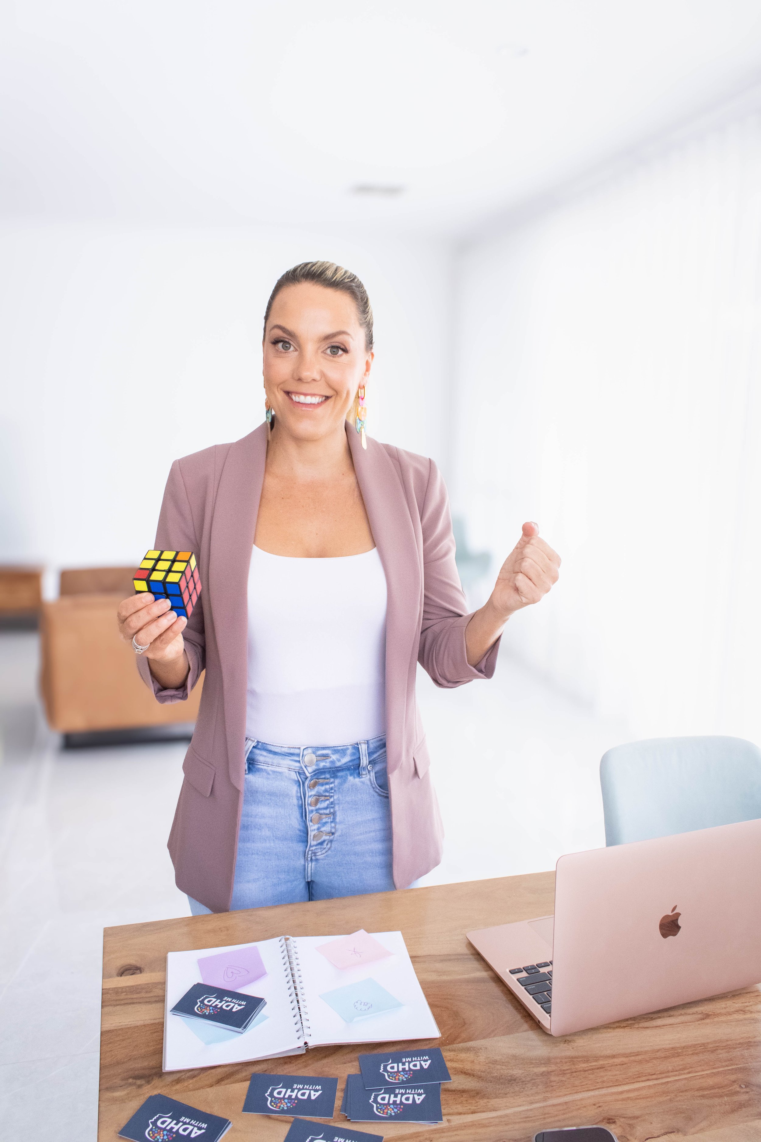Eliza Dadson, ADHD With Me Founder,  smiling while holding a Rubik's cube and a clenched fist in a bright, modern office space with a wooden desk, a laptop, and colorful cards and papers.