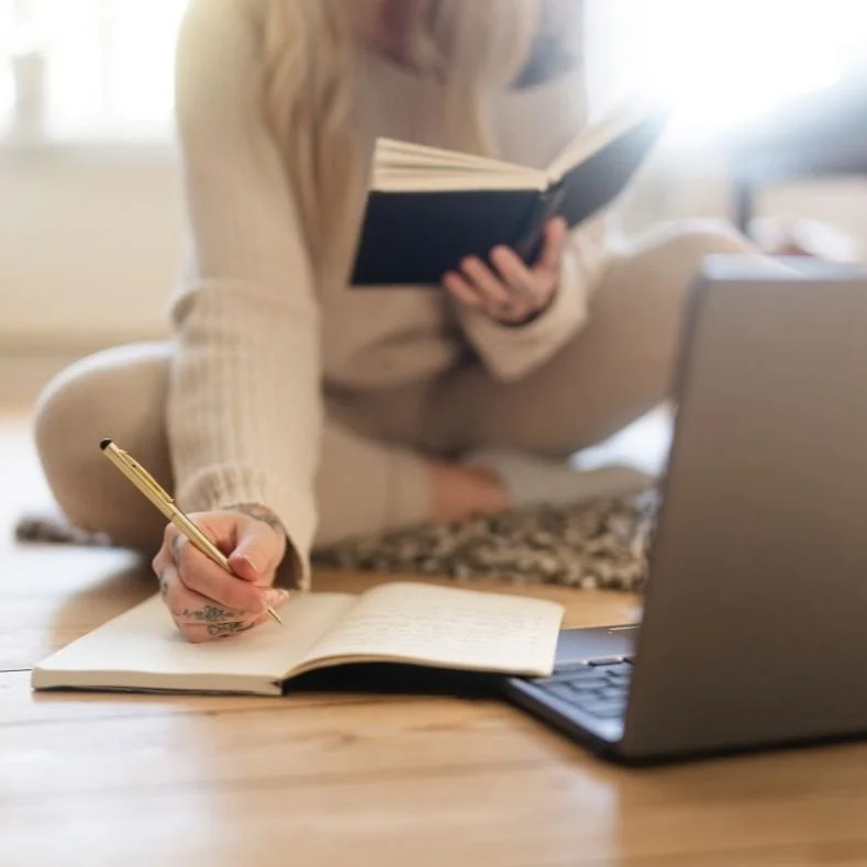 Person sitting on the floor with legs crossed, writing in a notebook, holding a book, with a laptop in front.