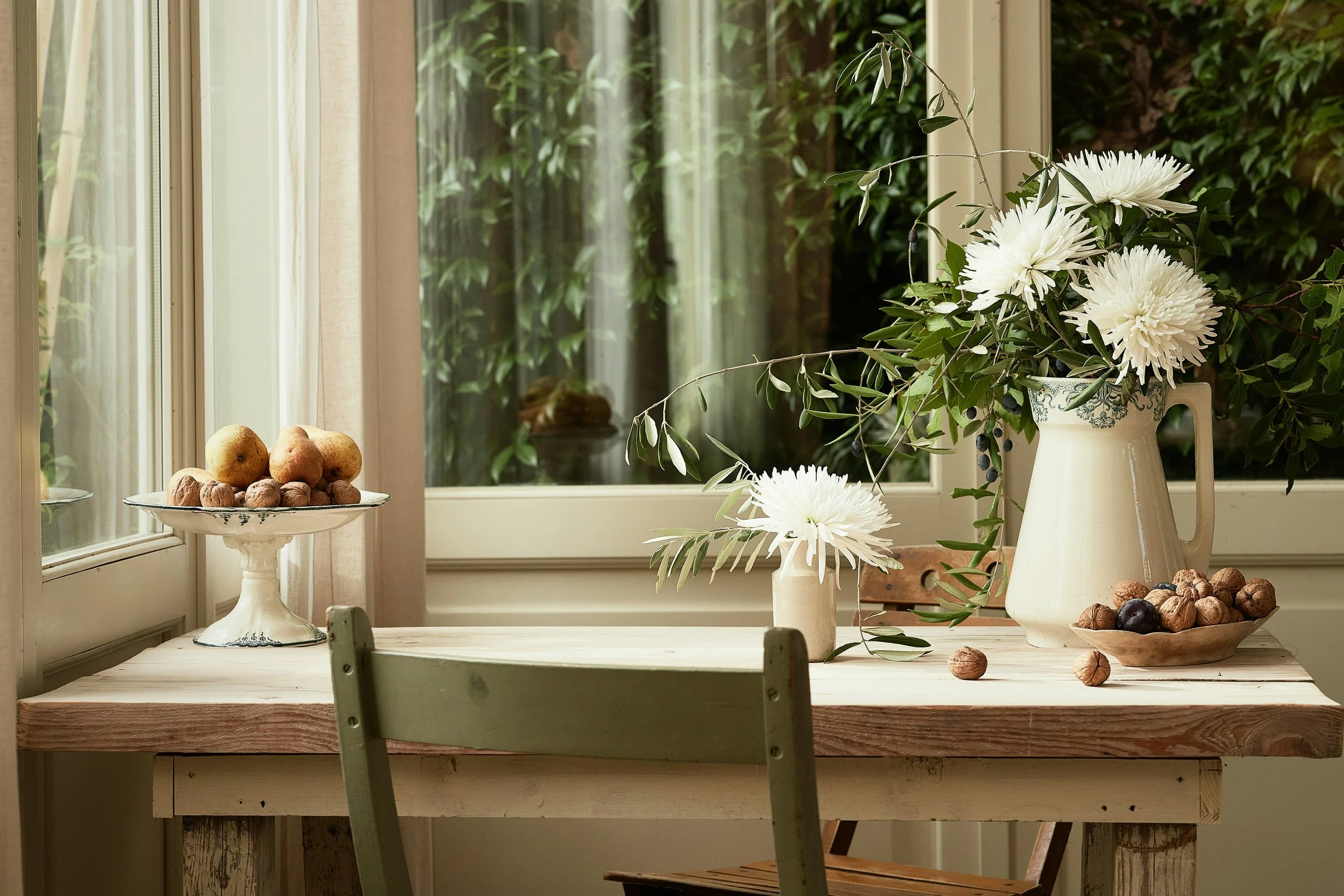 A wooden dining table with white floral arrangements in vases and a bowl of nuts, positioned near a window with greenery outside.