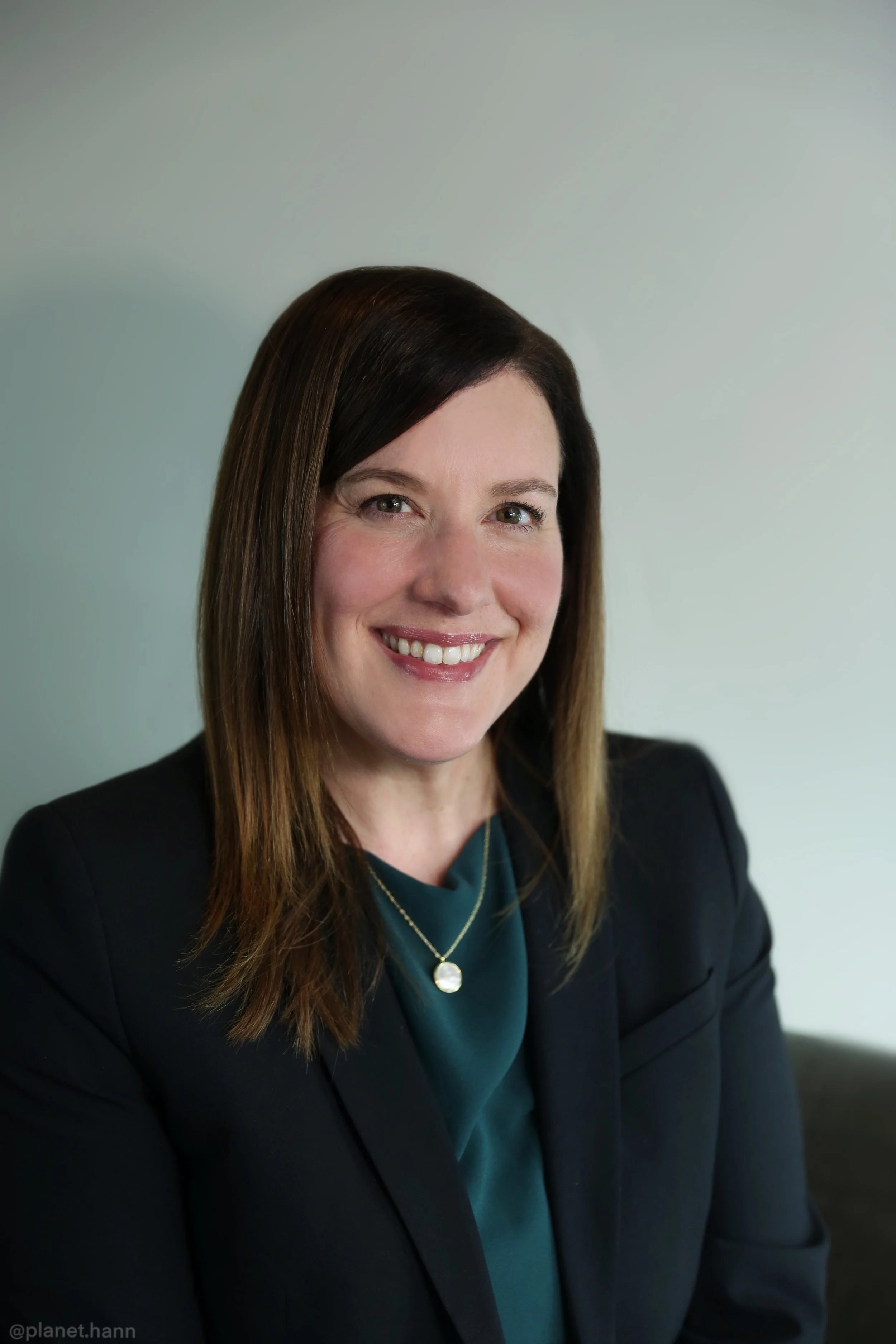 A woman with shoulder-length brown hair, wearing a black blazer and teal blouse, smiling at the camera against a plain light background.