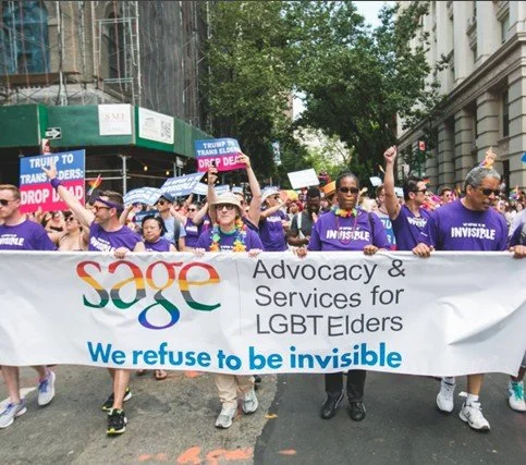 Group of people marching in a parade holding a large banner that reads 'SAGE Advocacy & Services for LGBT Elders. We refuse to be invisible' with some participants holding signs advocating for transgender rights.