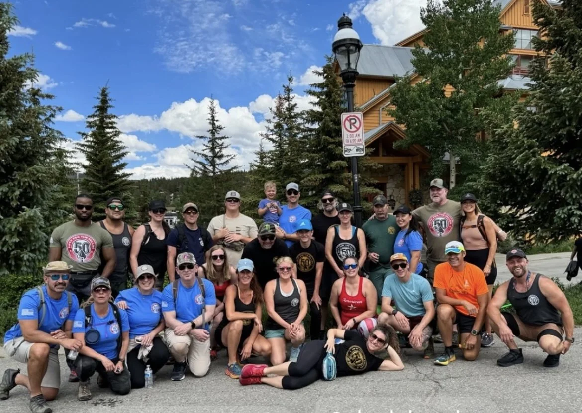 A group of people posing outdoors in front of trees and a building, some wearing matching shirts, with a clear sky above.