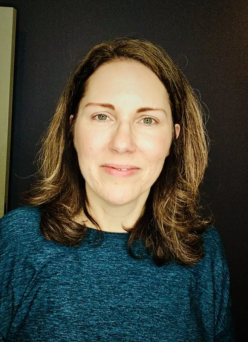 A woman with shoulder-length brown hair and light skin, wearing a blue textured top, standing in front of a dark background.