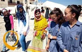 Group of women and girls outdoors at a festival or community event, with tents and a white canopy in the background. One woman is holding a yellow cloth, and some are wearing sunnies, smiling and engaging with each other.
